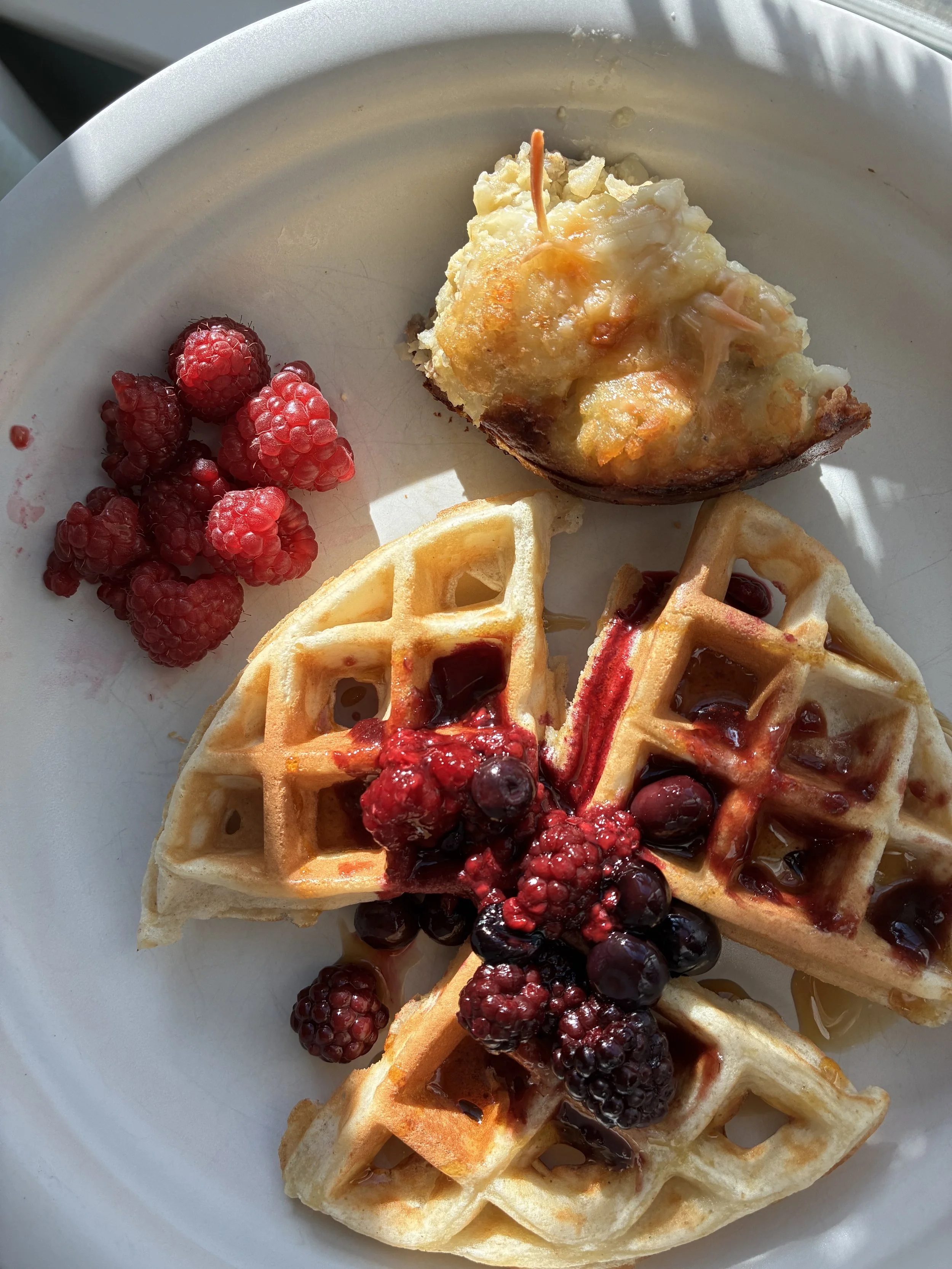 Waffles topped with mixed berries and berry syrup, served with a side of raspberries and a baked dish with cheese and vegetables.