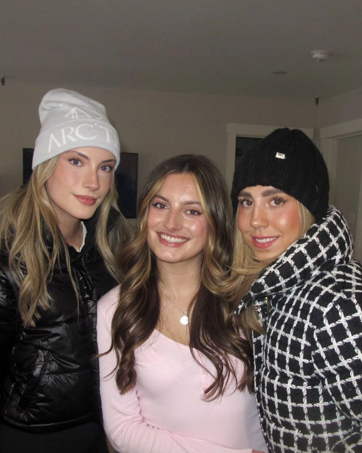 Three young women indoors, smiling, wearing winter clothing and hats.