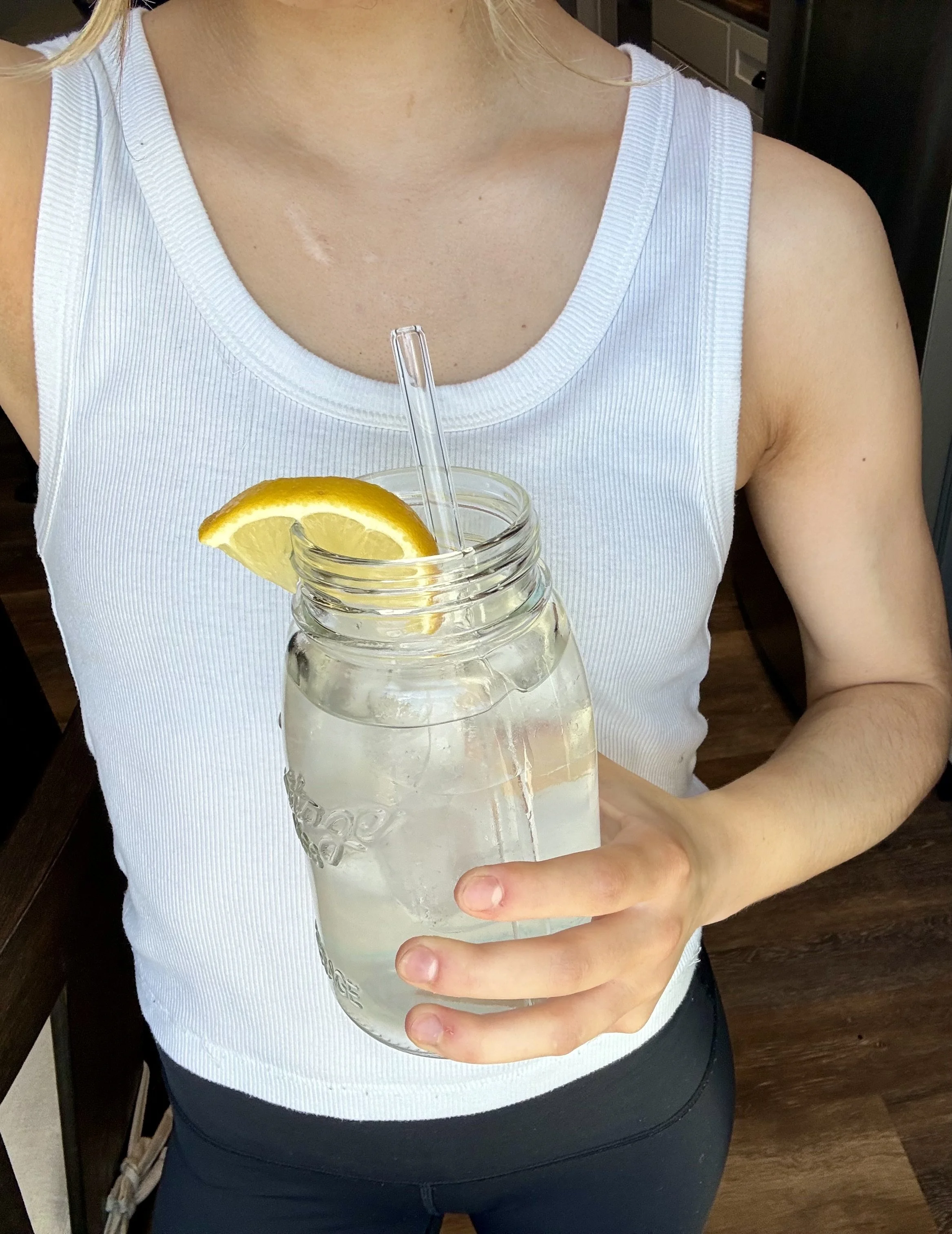 A person in a white tank top holding a mason jar filled with lemonade and ice, garnished with a lemon wedge, with a glass straw inside.