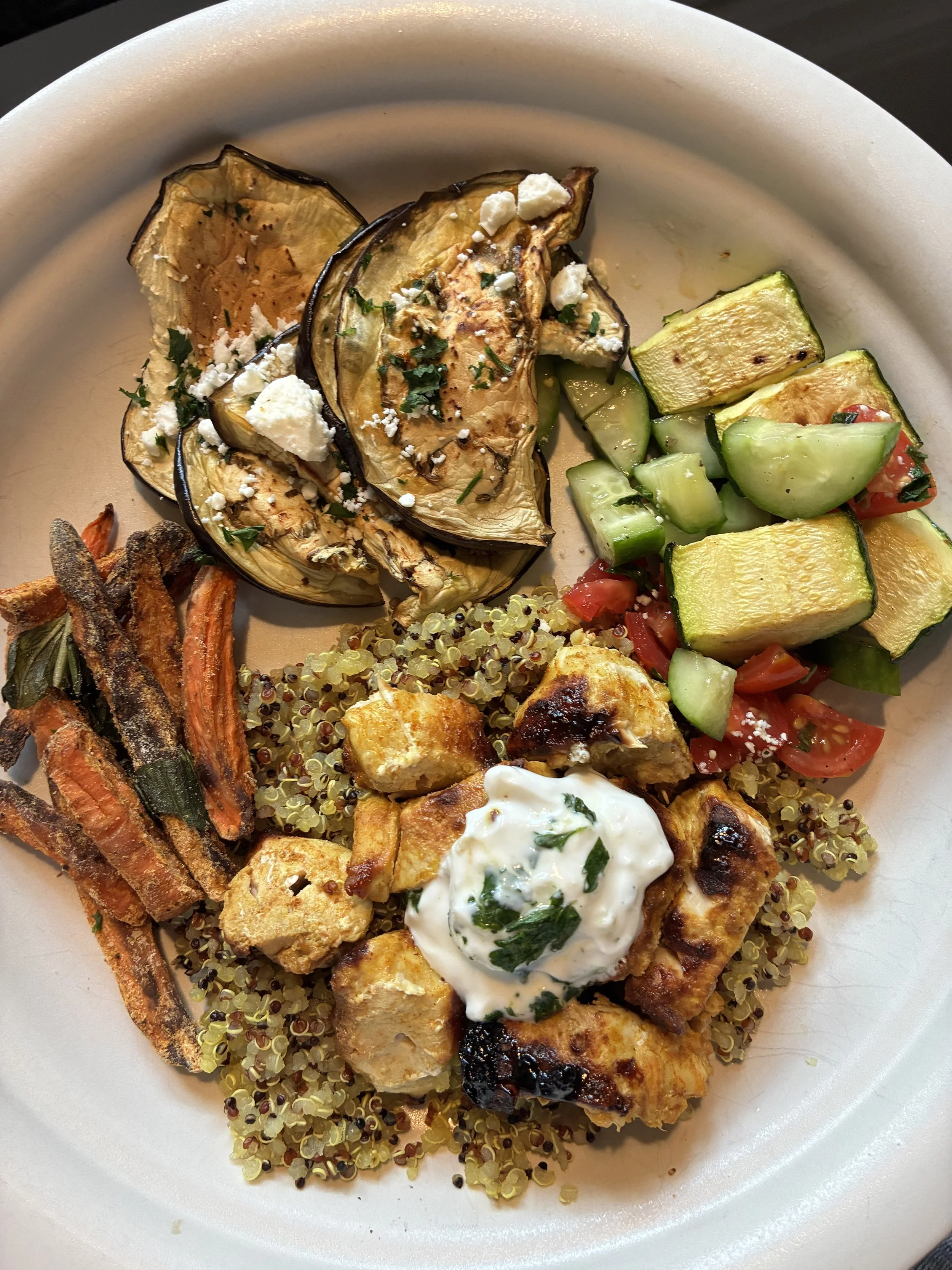 A plate of grilled eggplant topped with crumbled cheese and herbs, roasted carrots, a quinoa salad with grilled chicken and a dollop of creamy sauce, and a side of chopped cucumber and tomato salad.