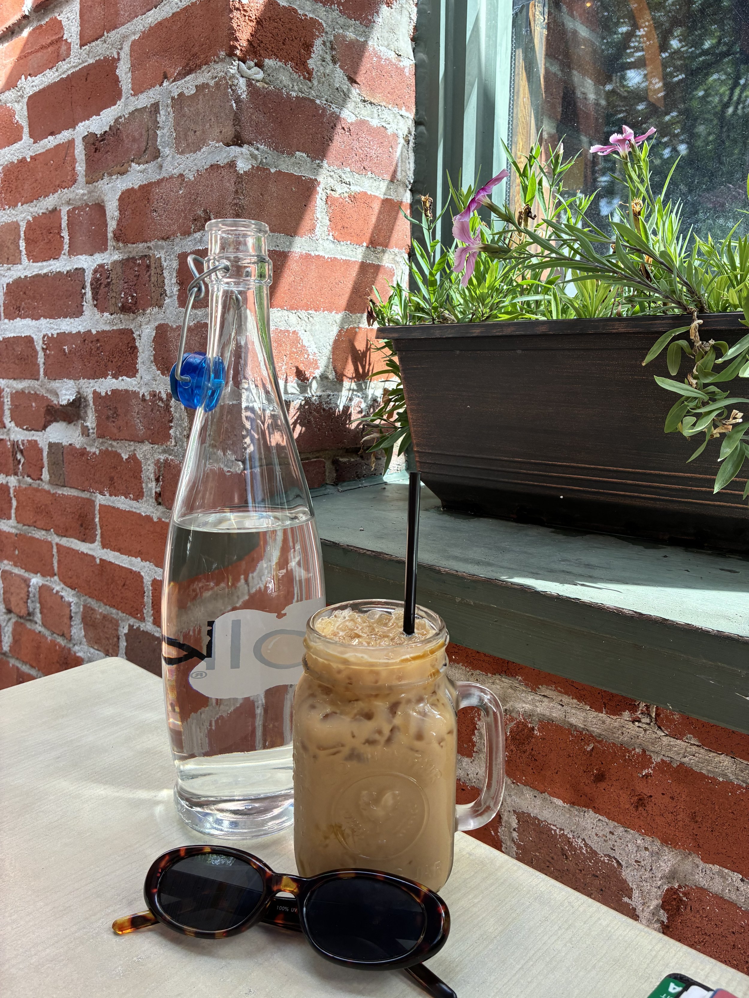 A glass bottle of water, a mason jar of iced coffee with a black straw, a pair of tortoiseshell sunglasses, and a small green and white object on a light-colored table near a brick wall and a windowsill with a potted flowering plant.