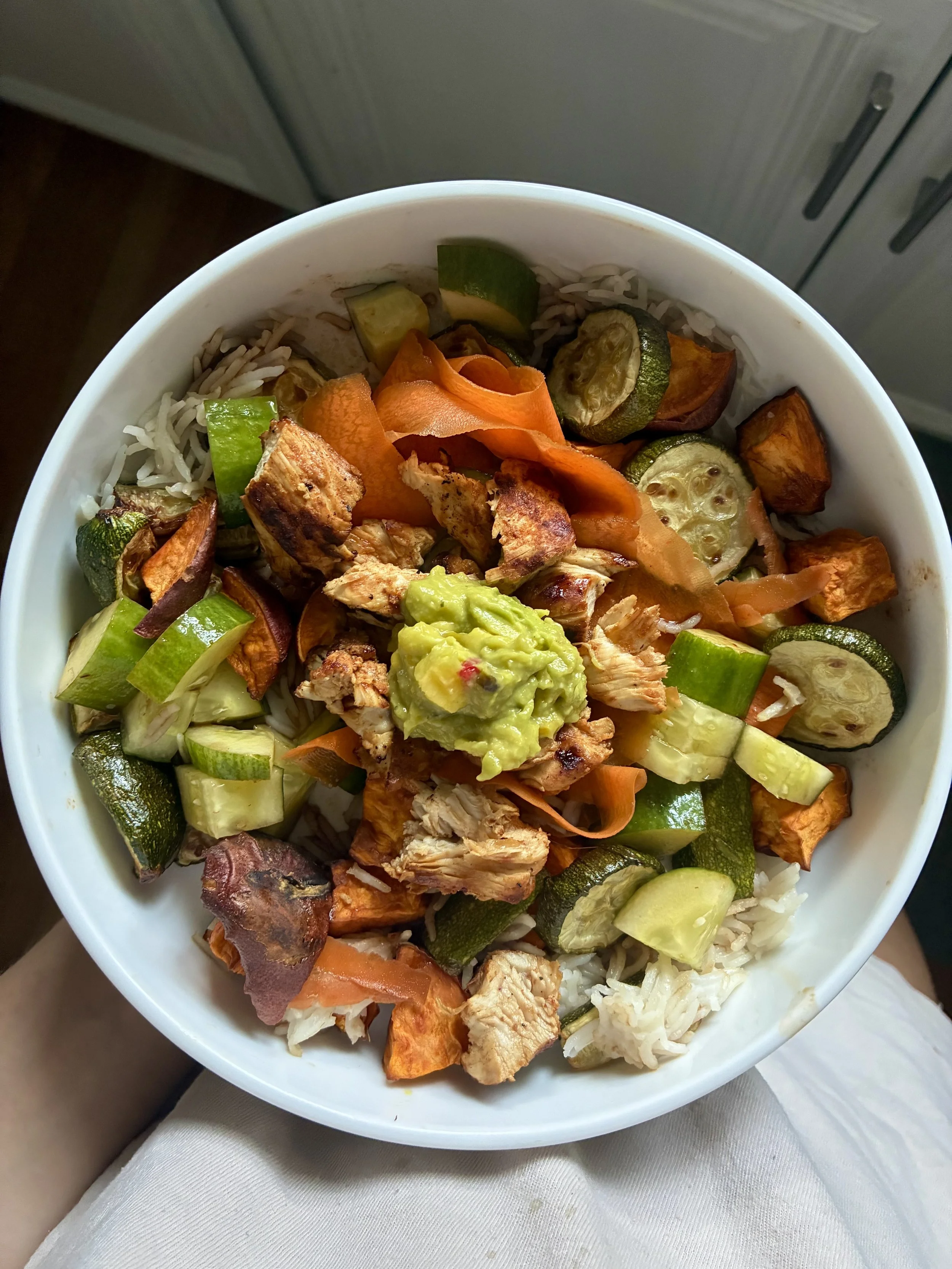A bowl of rice topped with grilled chicken, sliced zucchini, carrots, sweet potatoes, and guacamole.