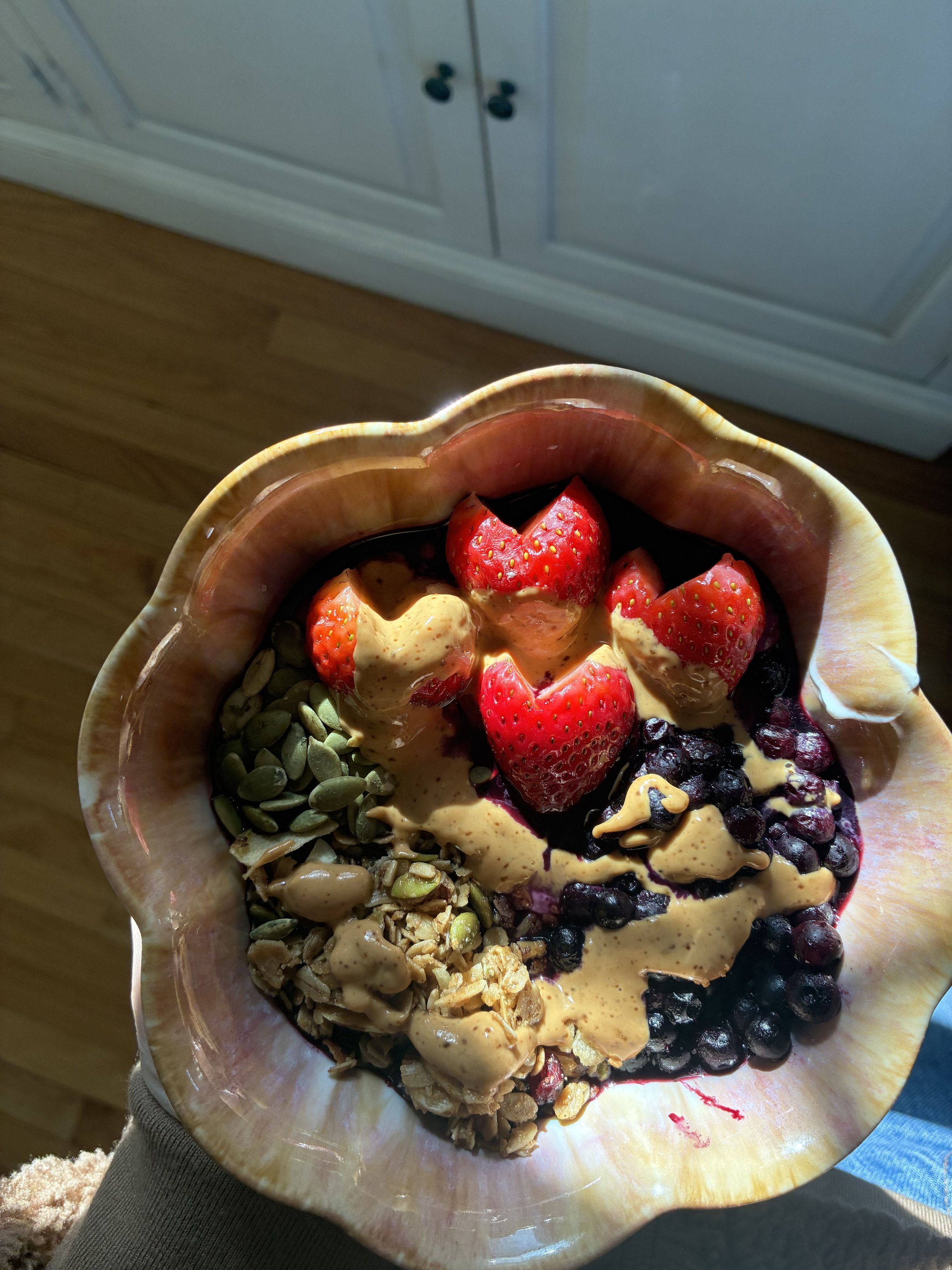A bowl of frozen yogurt topped with strawberries, blueberries, pumpkin seeds, granola, and peanut butter, in a bowl with a shell-like shape.