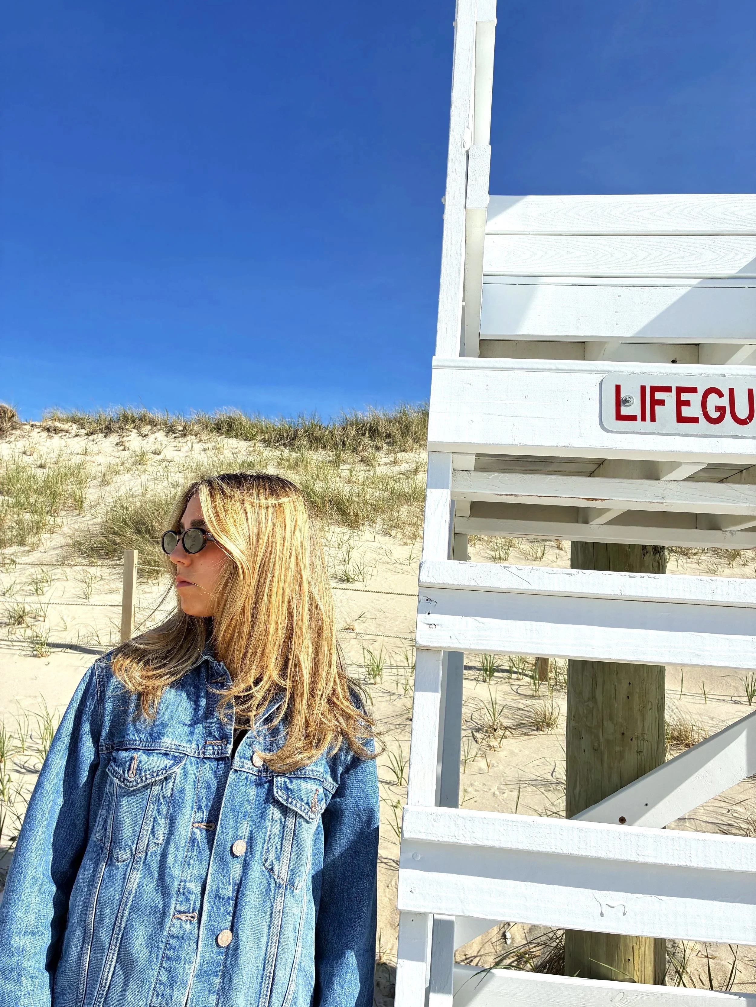 A woman with blonde hair wearing sunglasses and a denim jacket standing on a sandy beach near a lifeguard stand with a clear blue sky in the background.