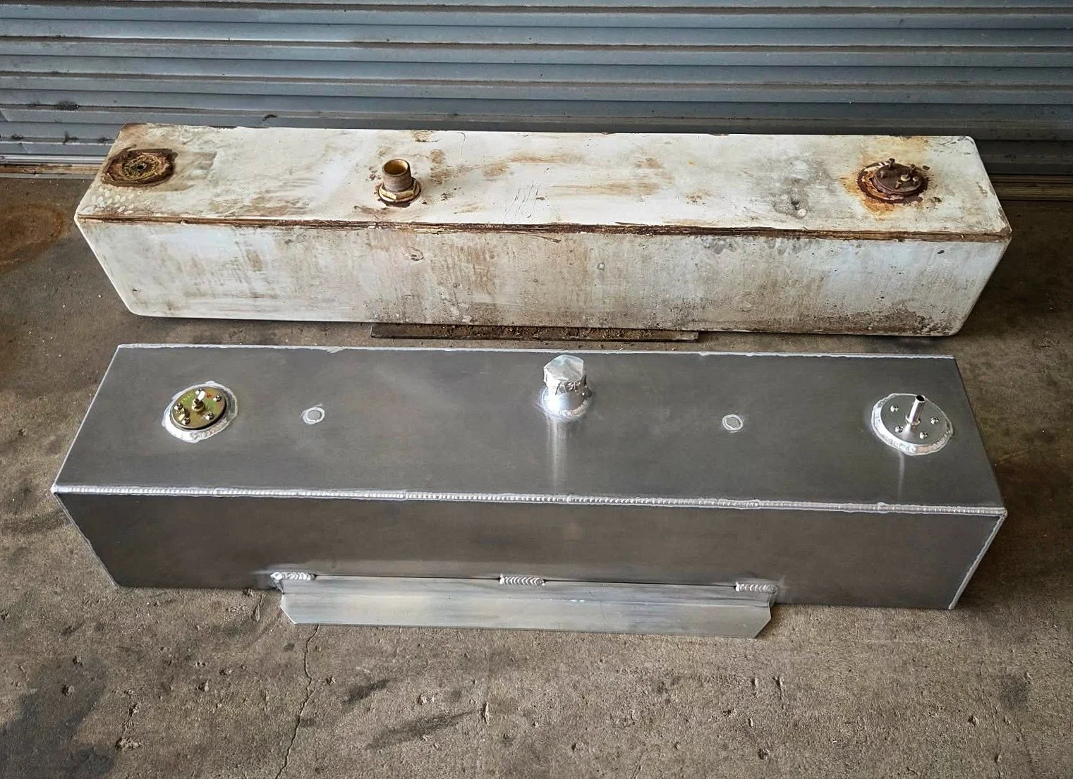 Comparison of two rectangular metal tanks, one old and rusty, the other new and shiny, placed on a concrete floor in front of a corrugated metal wall.