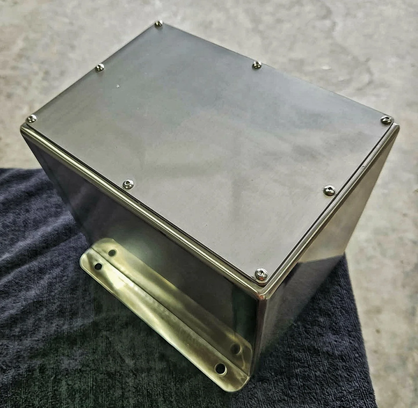 A rectangular metal enclosure with a flat top, secured with screws at each corner, held in a person's hand against a concrete surface background.