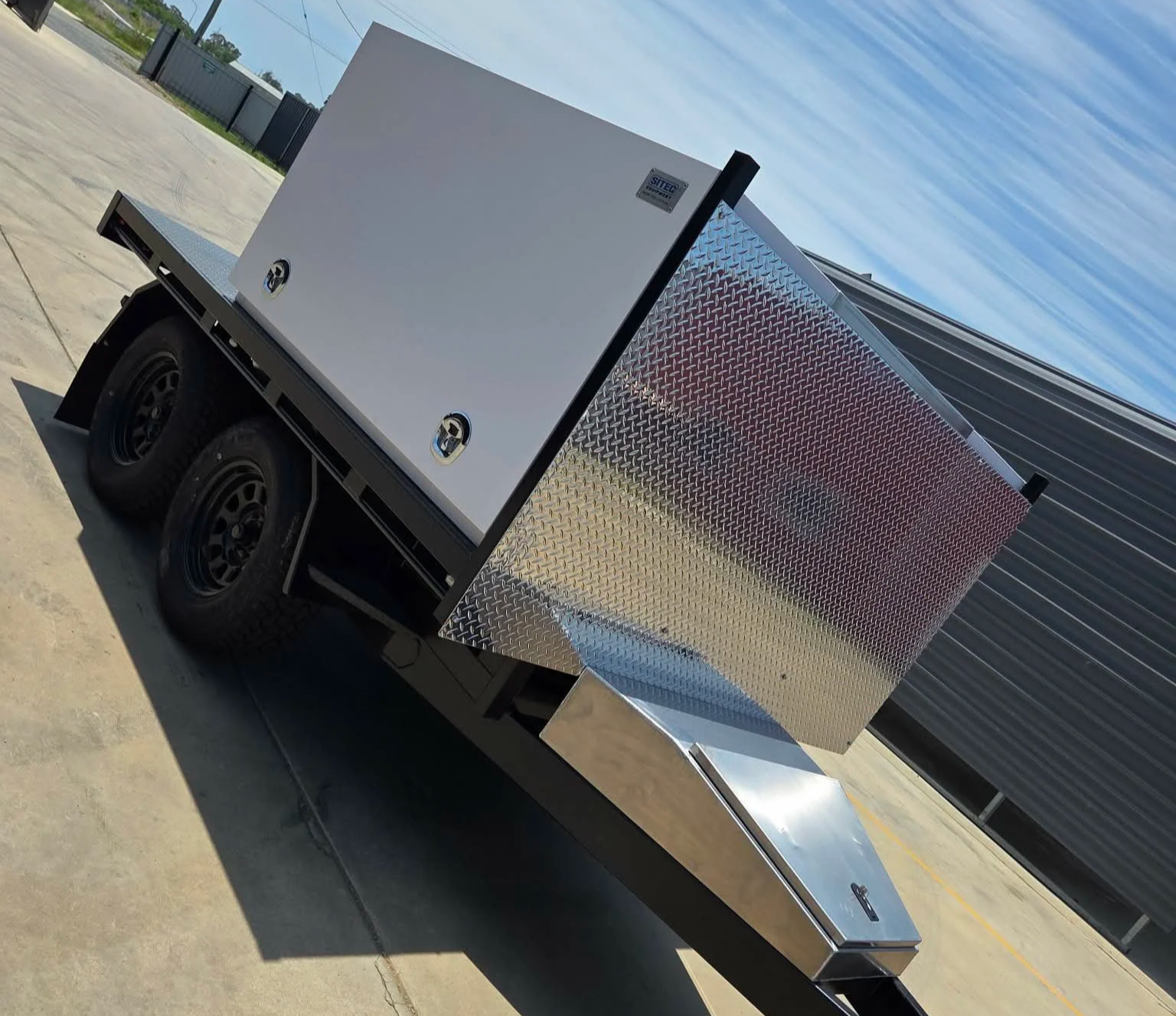 A utility trailer with two tires, a white and gray body, and a metallic storage box mounted on it, parked on a concrete surface outside under a clear sky.