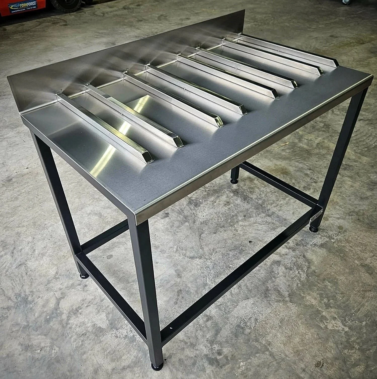 Stainless steel table with built-in drainage or chute, supported by black metal frame, in a warehouse or workshop setting.