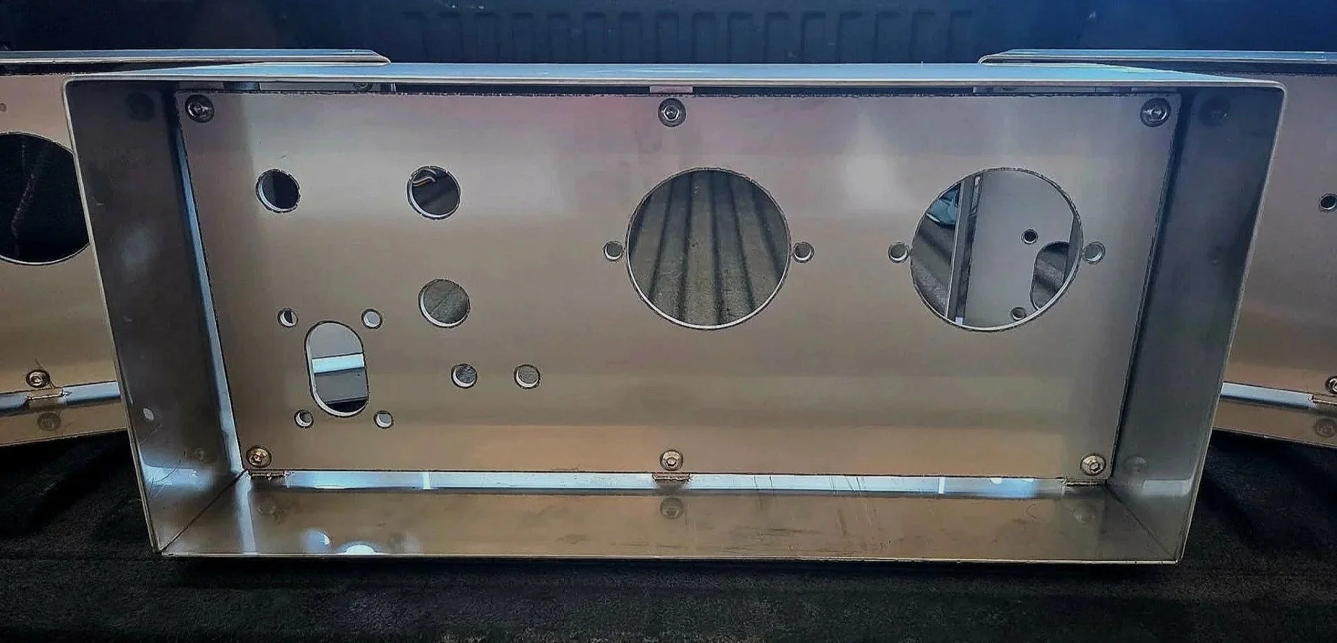 A metallic rectangular electronic enclosure or chassis with multiple circular and oval holes, placed on a black surface.