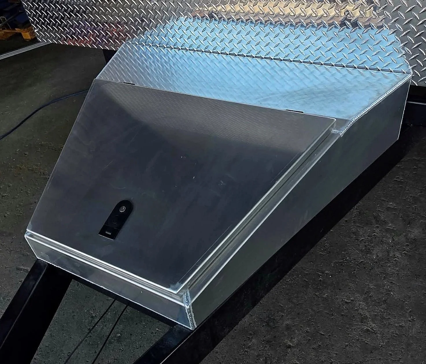 Metal utility trailer with diamond plate surface, black handle, and electronic lock, parked on a dark concrete surface.