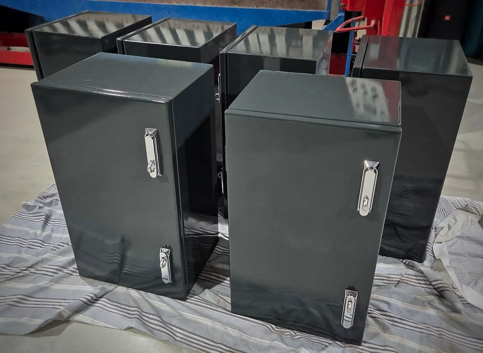 Five gray metal storage cabinets with silver handles arranged on a striped cloth. The cabinets have a polished finish and are positioned on a concrete floor in an indoor setting.