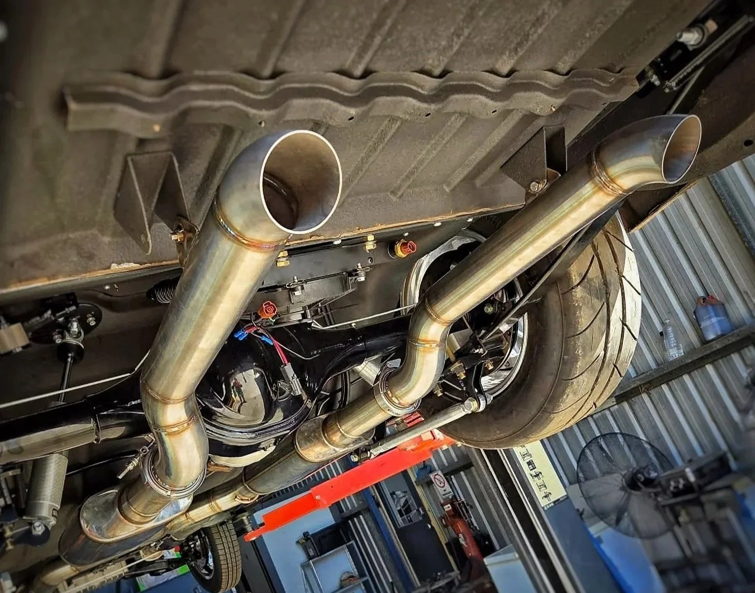 Underside of a car showing custom exhaust pipes, rear tire, and parts of the suspension and chassis in a garage or workshop.