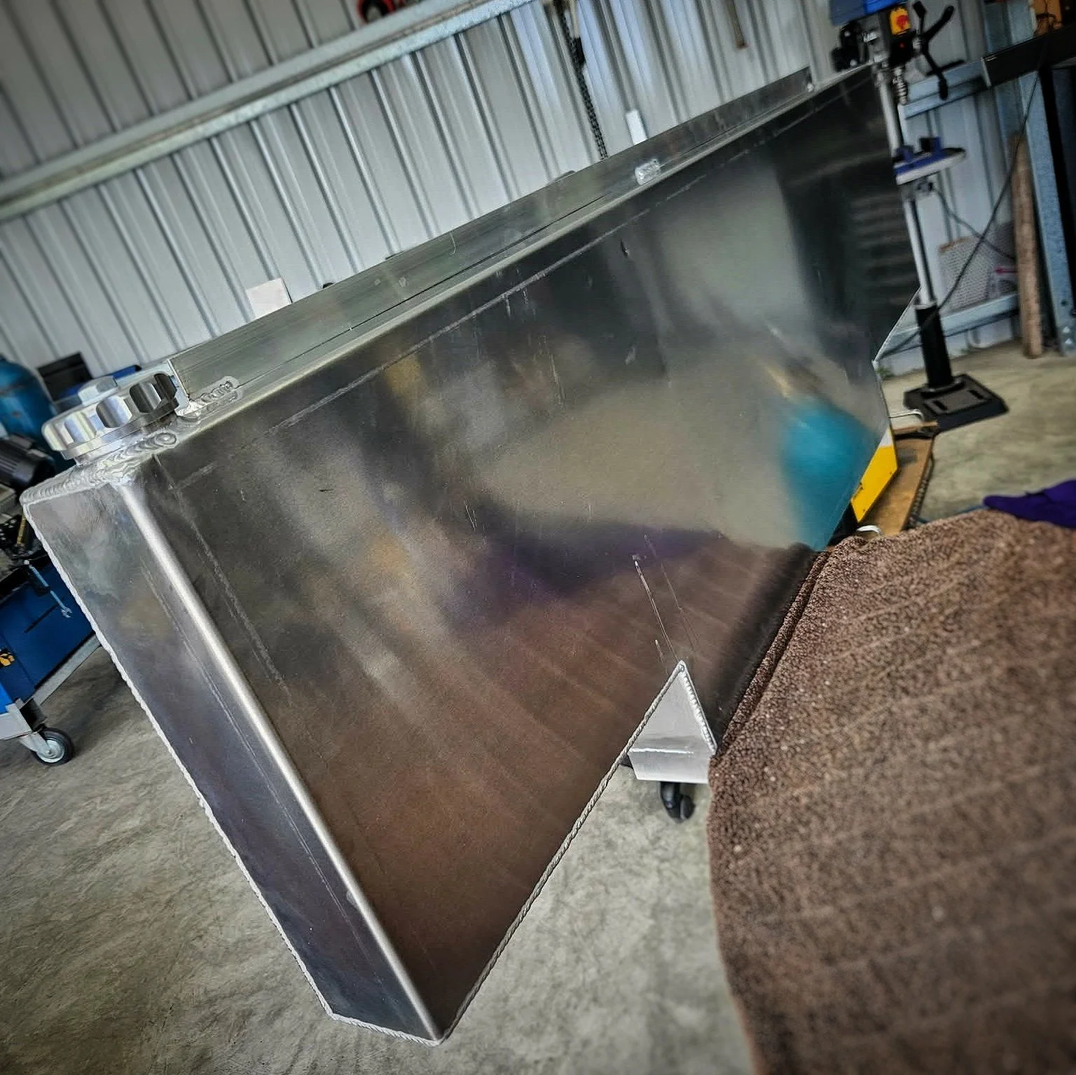 A large stainless steel industrial equipment or appliance in a workshop or garage setting, with a textured brown towel or cloth nearby