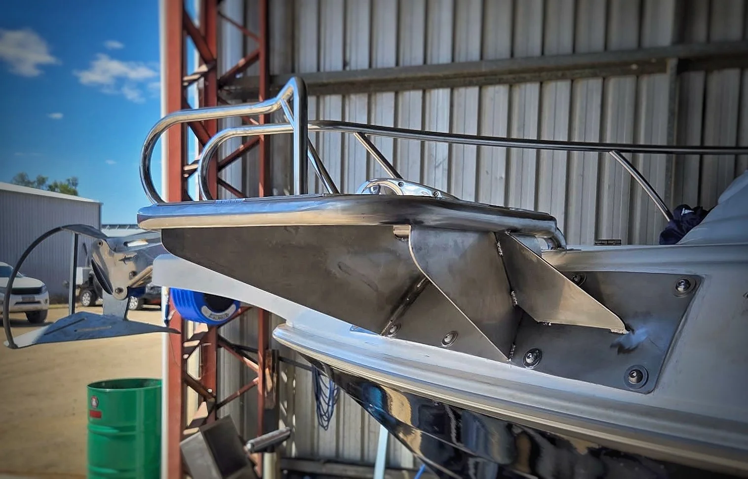 Close-up of a metallic boat hull under construction or repair, with a metal framework and part of a boat's side visible, outdoors with a blue sky and some buildings in the background.