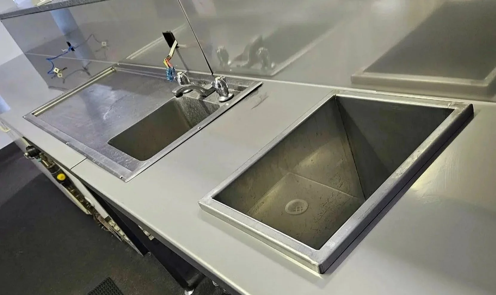 Stainless steel commercial kitchen workstation with a sink, an empty slot for a stove or appliance, and another empty compartment, with wiring and plumbing visible.