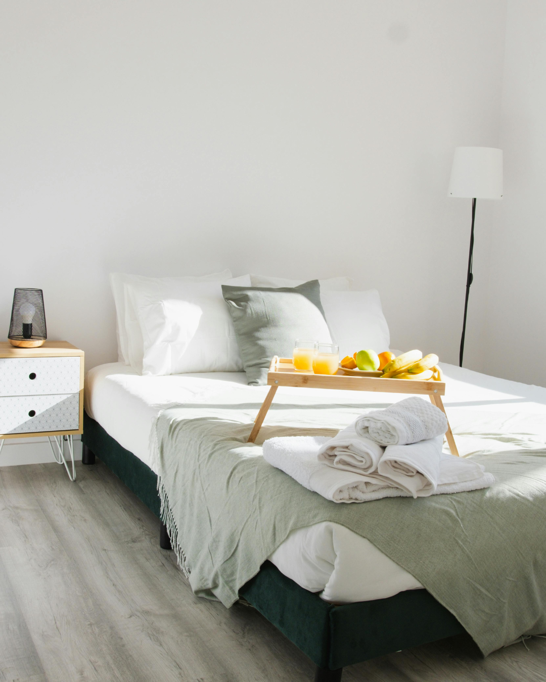 A neatly made bed with white linens and pillows in a bright, minimalist bedroom. A wooden breakfast tray with orange juice and fruit is on the bed, along with folded towels.