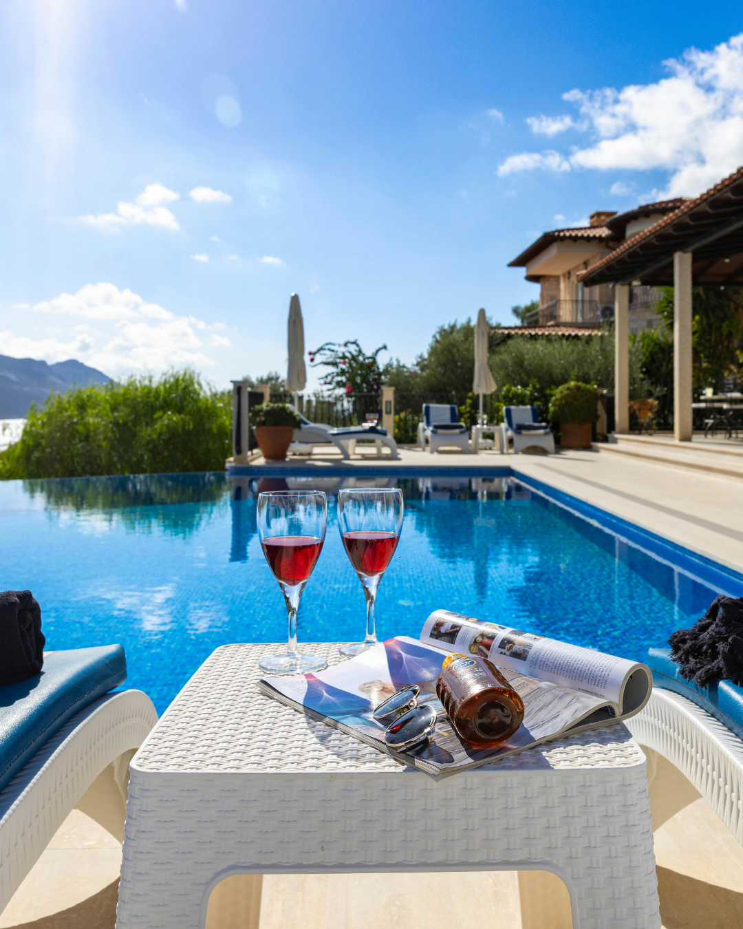 Poolside scene with two glasses of red wine, sunglasses, a bottle, and an open magazine on a white wicker table, overlooking a blue swimming pool, lounge chairs, umbrellas, and a house under a bright blue sky.