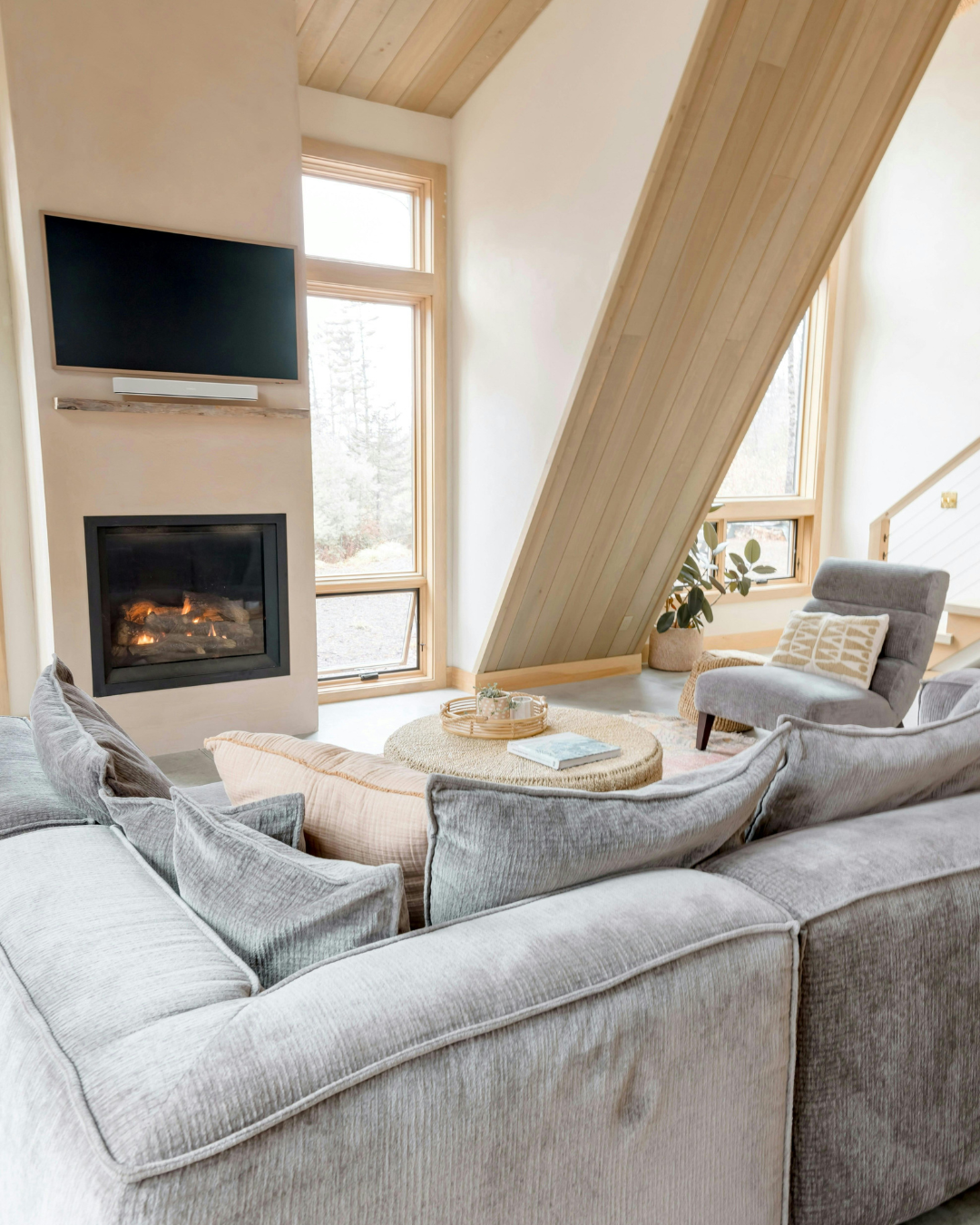 Cozy living room with a gray sectional sofa, a round wicker coffee table with a book and tray, a gray armchair with a cushion, a large plant, and windows with natural wood trim. There is a fireplace and a wall-mounted TV.