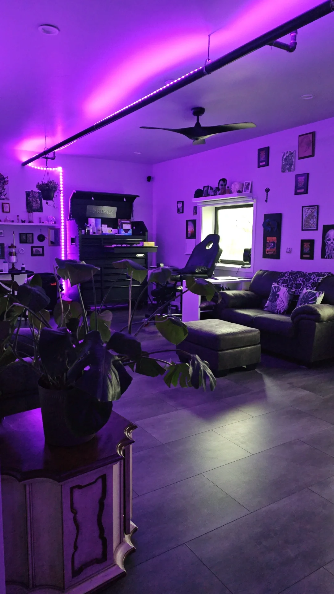 Living room illuminated with purple LED strip lights, featuring a black piano with various items on top, a black reclining chair, a black sofa with patterned pillows, framed art on the walls, a window with natural light, and a houseplant in the foreground.