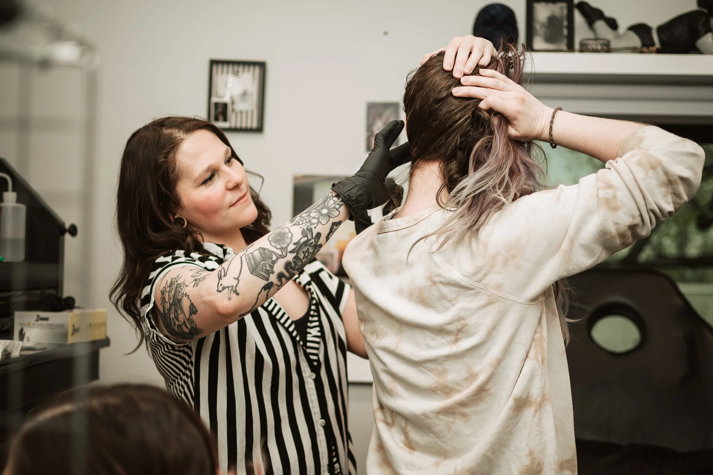 Tattooed woman with black gloves helping a woman with dyed hair in a tattoo shop.