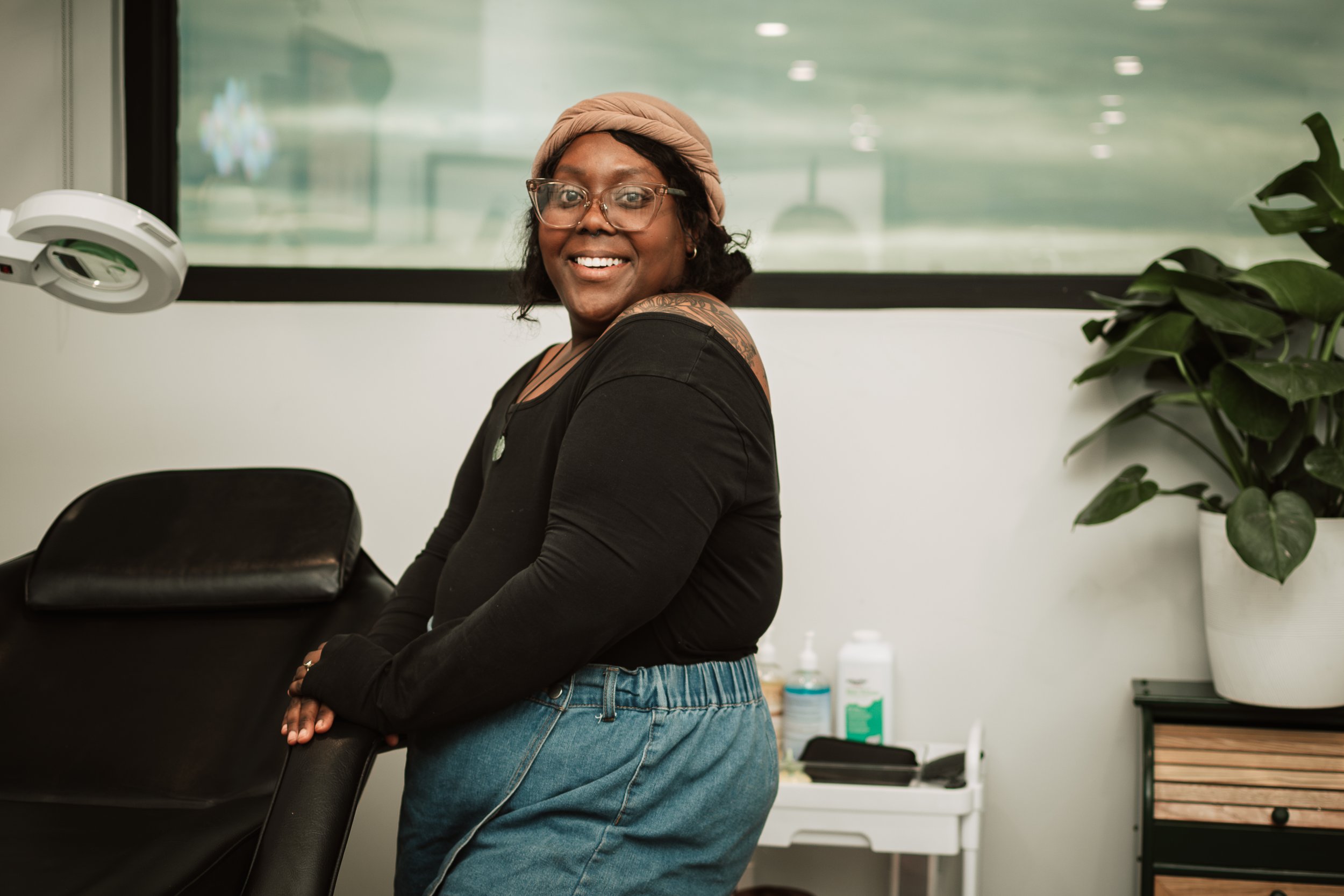 A smiling woman with glasses, wearing a tan headwrap, black long-sleeve top, and blue jeans, is standing in a medical or beauty clinic. She is leaning on a black chair and appears to be relaxed and happy. There is a large plant in a white pot and medical supplies on a countertop in the background.