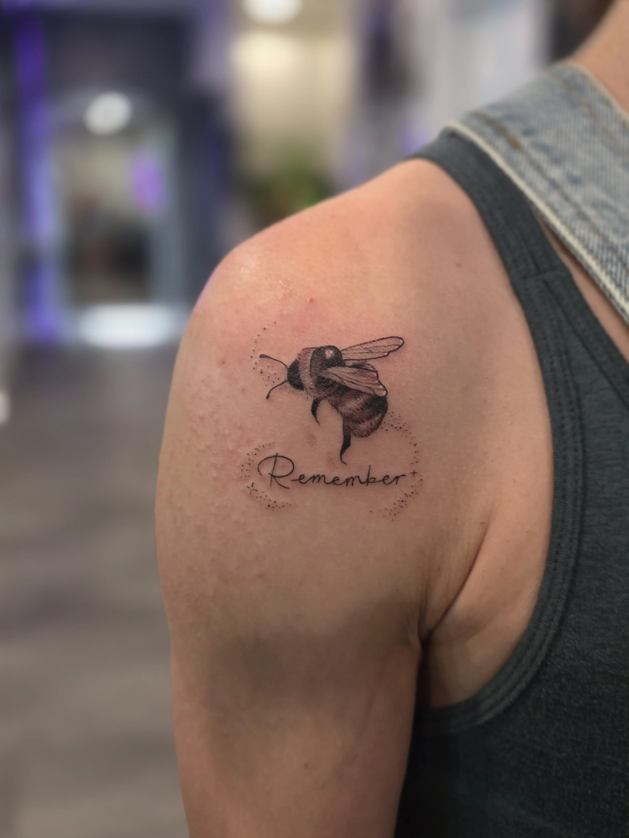 Tattoo of a bee with the word 'Remember' written underneath on a person's shoulder.