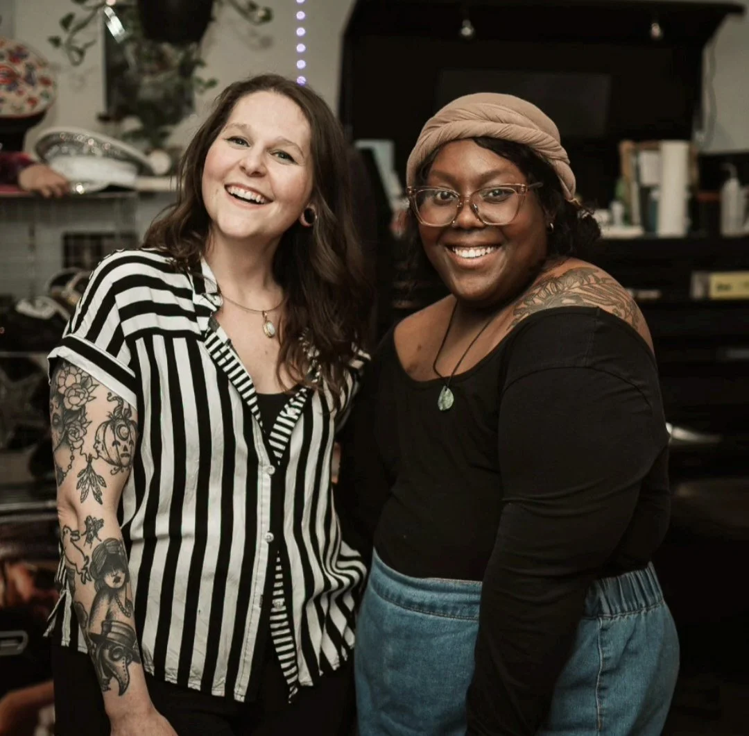 Two women smiling and standing close together in an indoor setting, one with tattoos on her arm wearing a black and white striped shirt, and the other wearing glasses, a beige head wrap, and a black top.