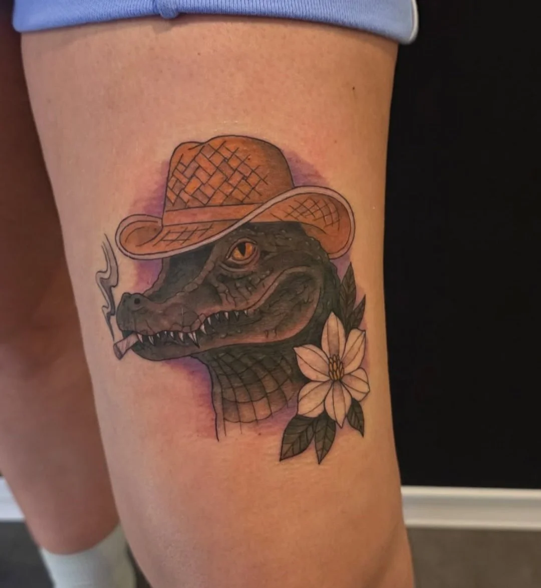 Tattoo of a dinosaur wearing a straw hat with a flower near its neck.