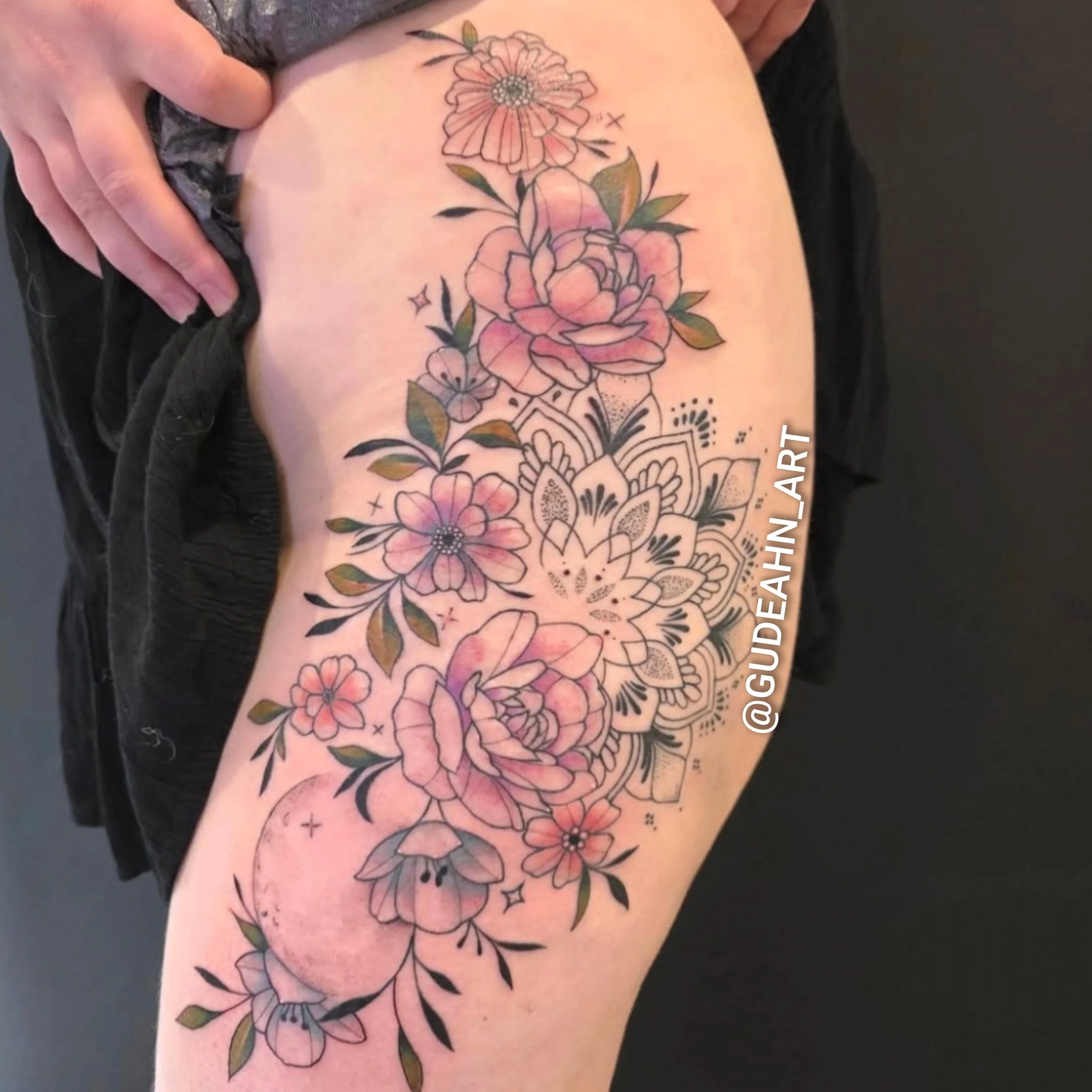 A colorful floral tattoo featuring various flowers with pink and purple petals, green leaves, and decorative black ink details on a person's thigh.