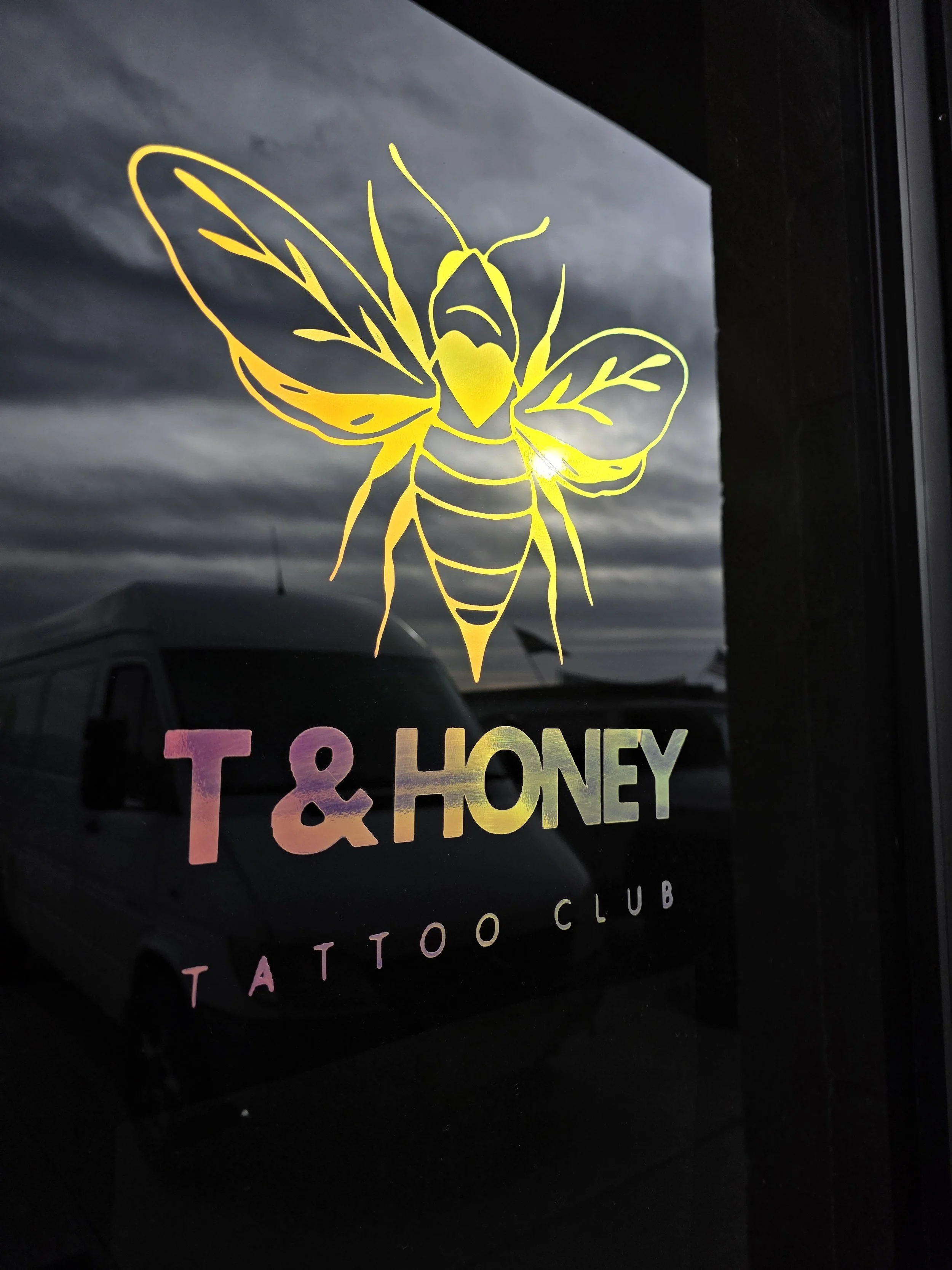 Sign on window for T & Honey Tattoo Club featuring a yellow outline of a bee with colorful text.