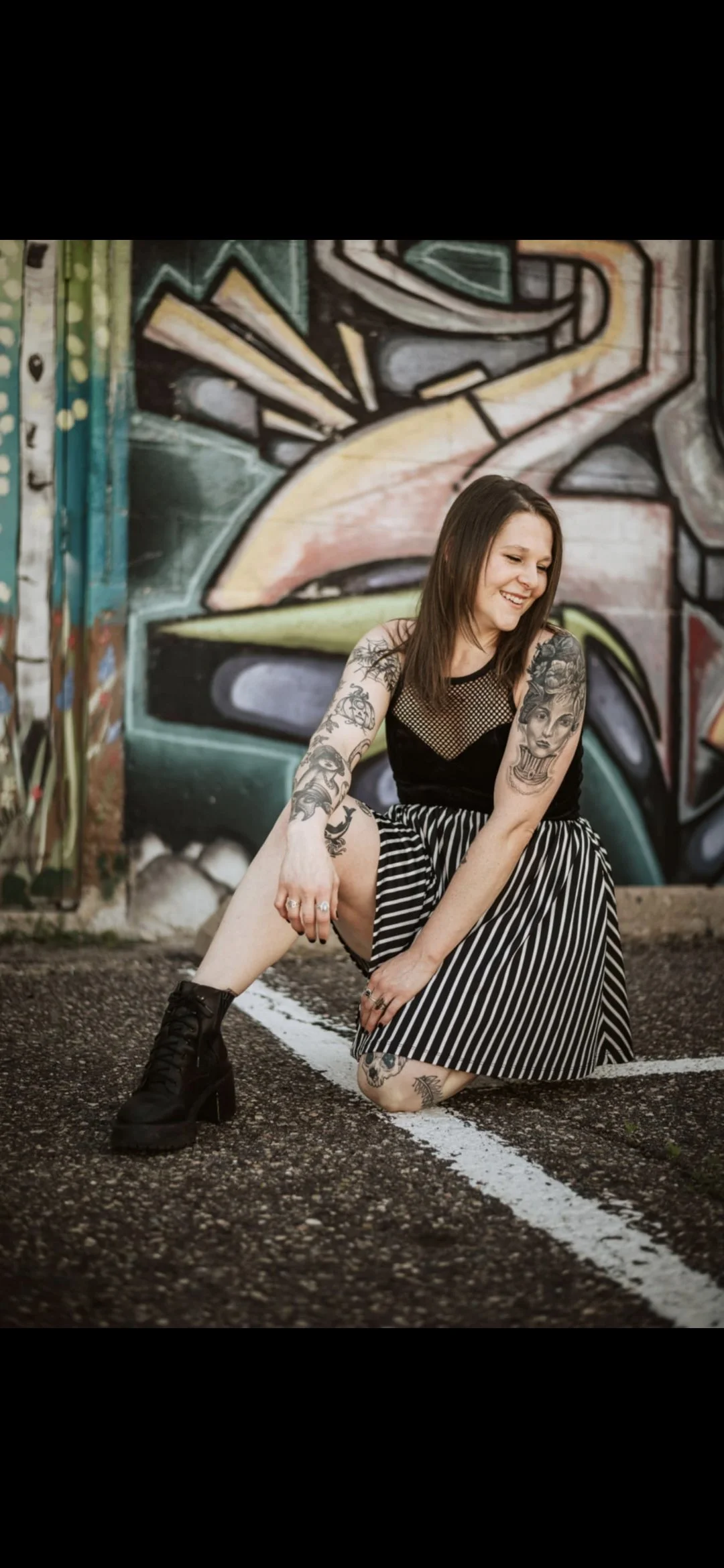 A woman with tattoos on her arms and legs, wearing a black and white striped skirt, black top, and black high-heeled boots, squatting on the ground and smiling near a graffiti-covered wall.