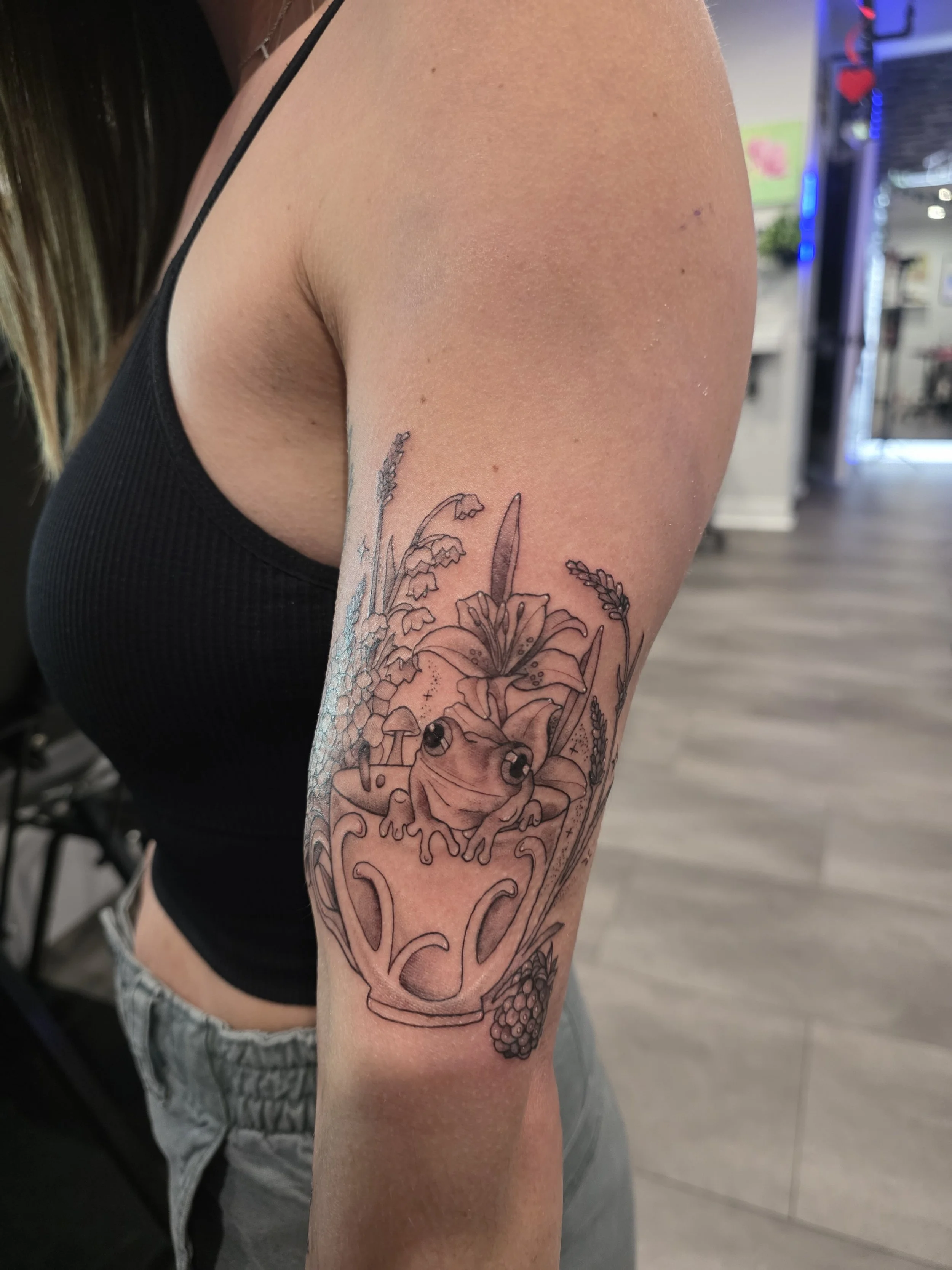 Tattoo of a frog inside a teacup with flowers, leaves, and small mushrooms surrounding it on a person's upper arm.