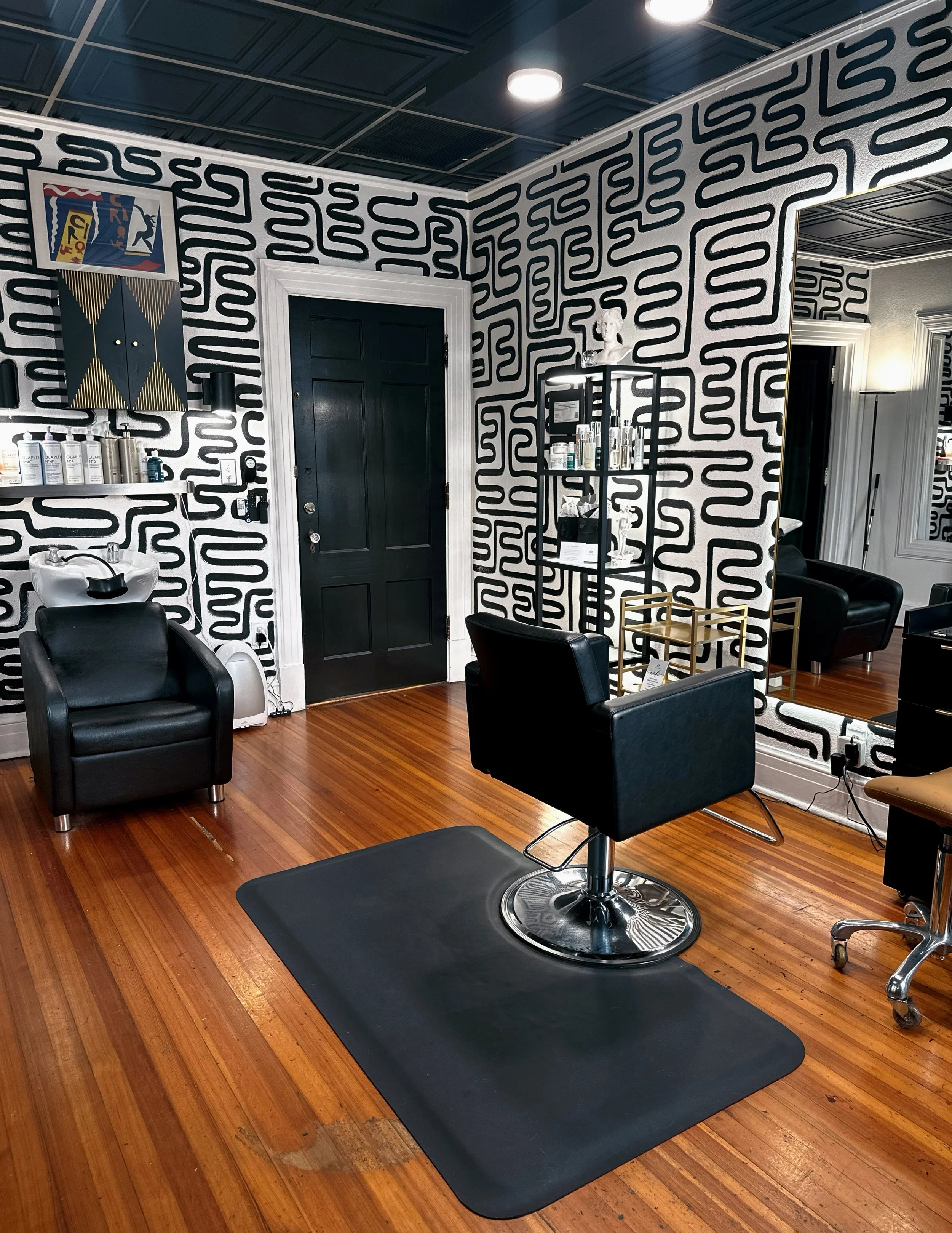 Barber shop with black and white patterned wallpaper, black door, styling chair on a black mat, sink, shelves with hair products, and a large mirror reflecting the room.