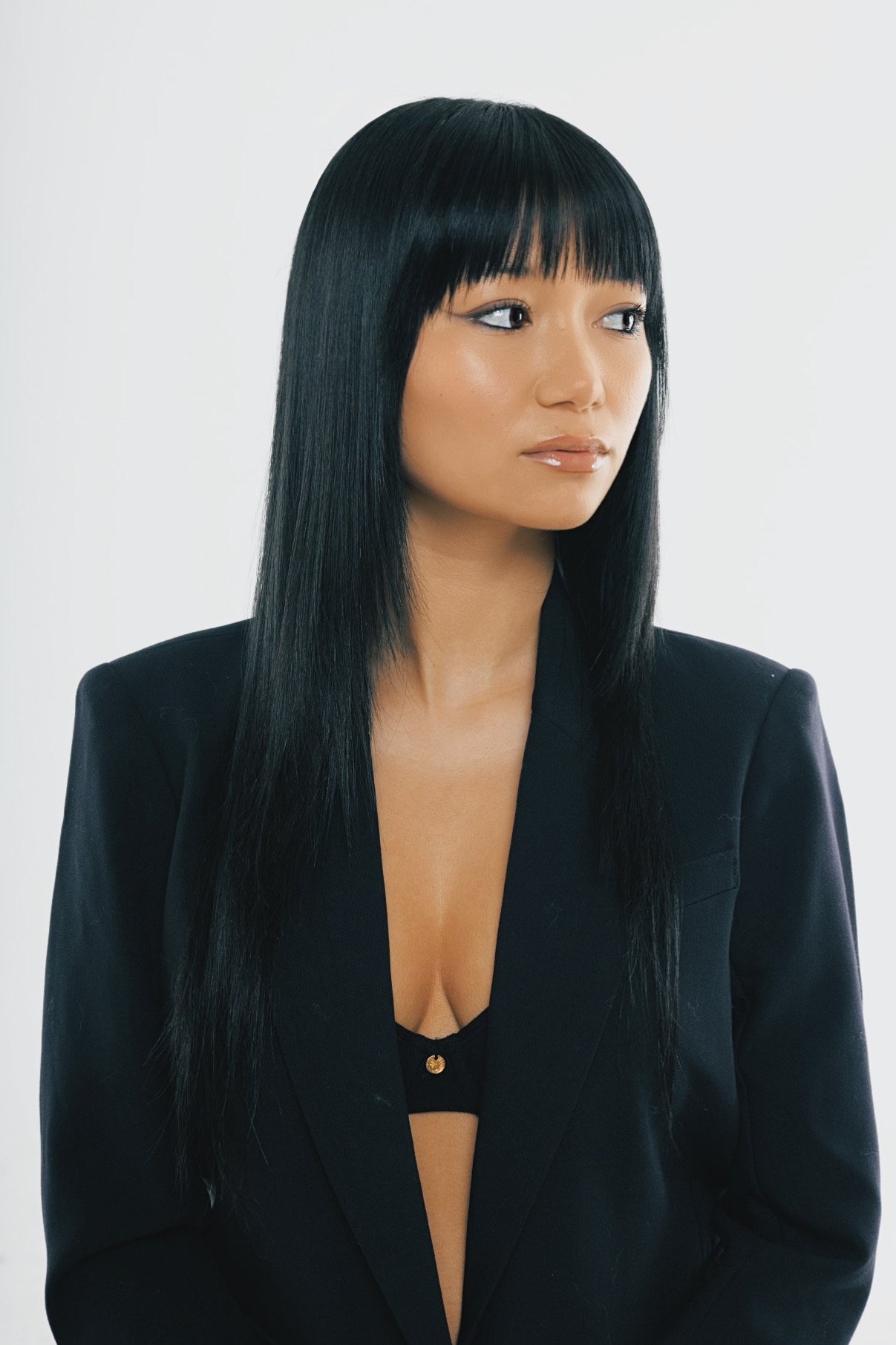 A woman with long black hair and bangs, wearing a black blazer and a black top with a low neckline, standing against a plain white background.