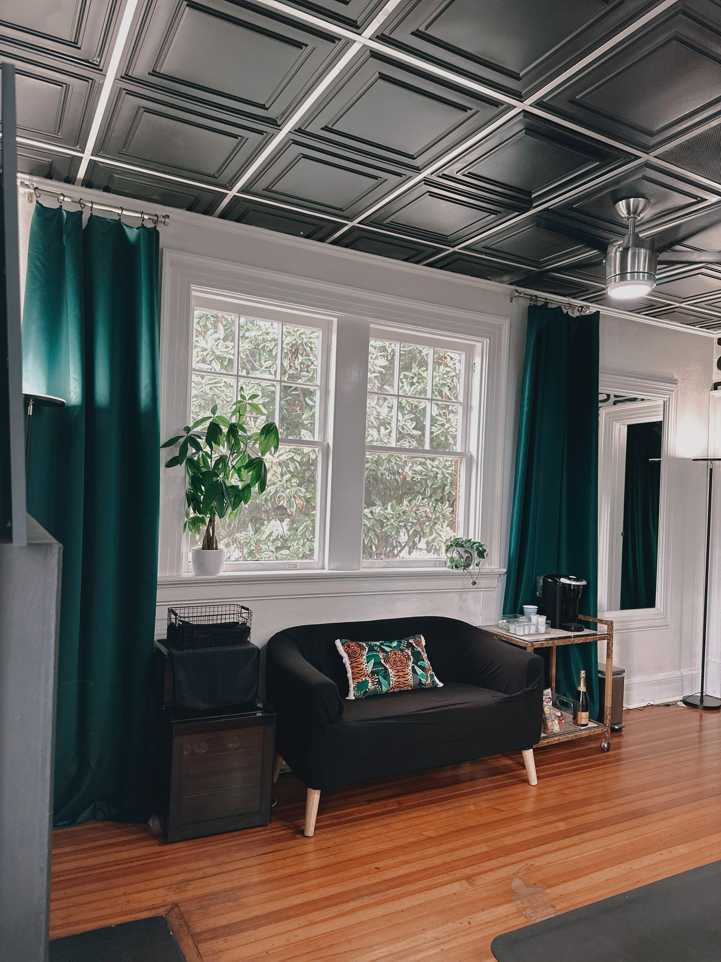 Living room with wooden floor, green curtains, a black armchair with a colorful animal print pillow, a small bar cart with drinks, a potted plant by the window, and a ceiling with black decorative panels.
