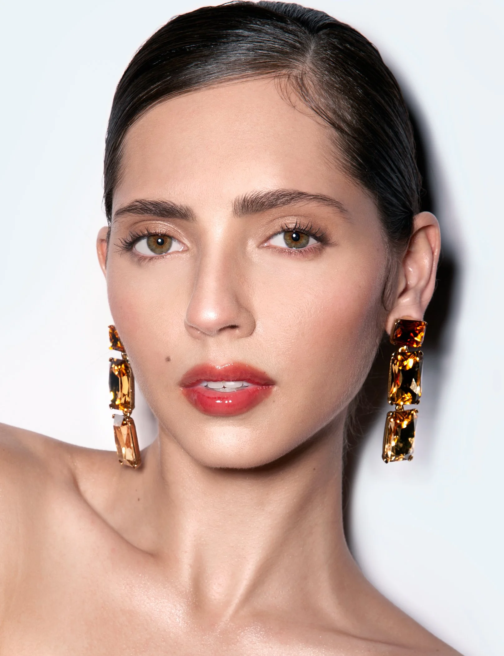 Close-up of a woman's face with dark, slicked-back hair, wearing large gold and amber earrings, with makeup including foundation, subtle eyeshadow, eyeliner, and pink lipstick, against a plain light background.