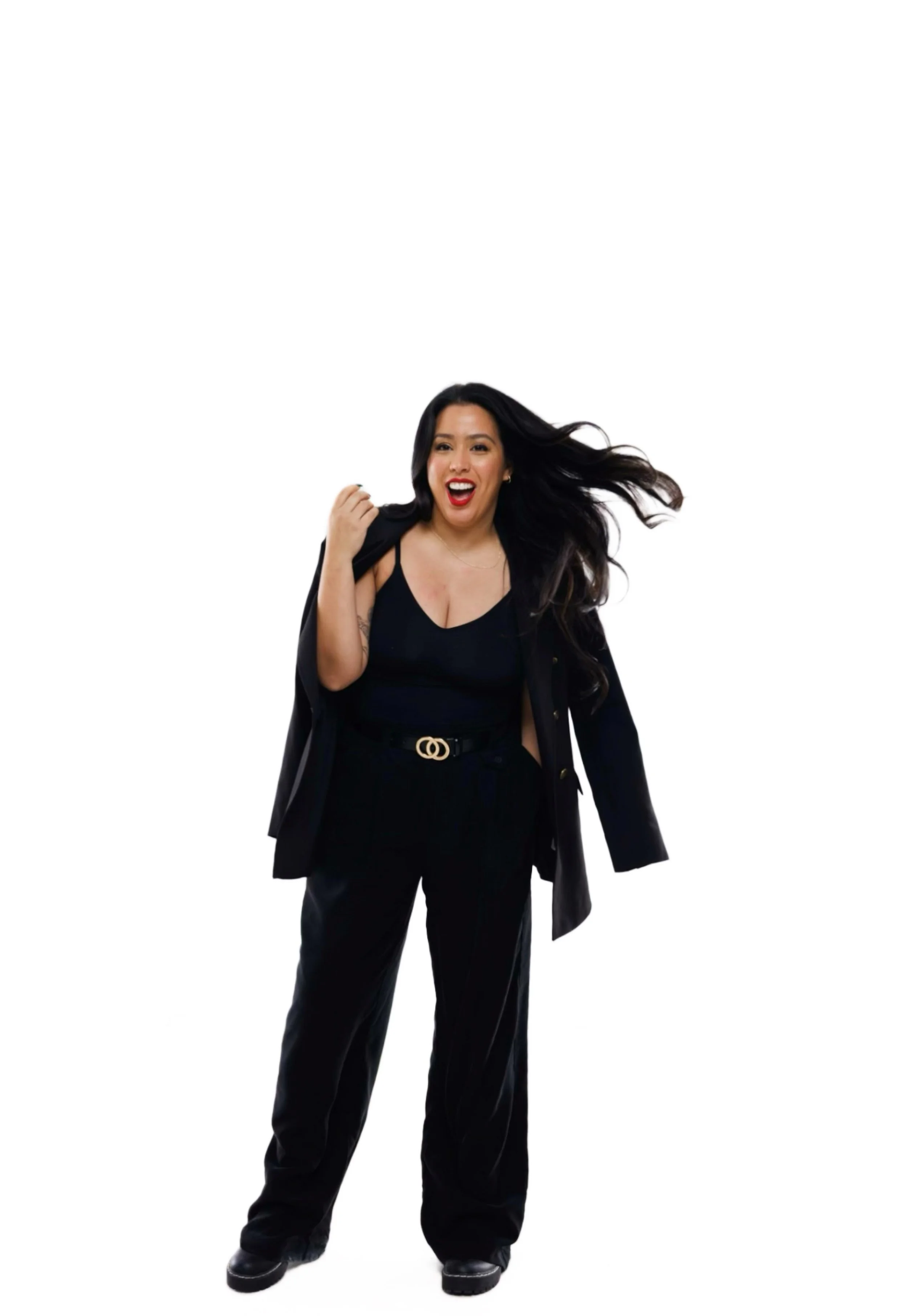 A woman with long dark hair, wearing a black sleeveless top, black pants, and a black blazer draped over her shoulders, smiling and posing confidently.