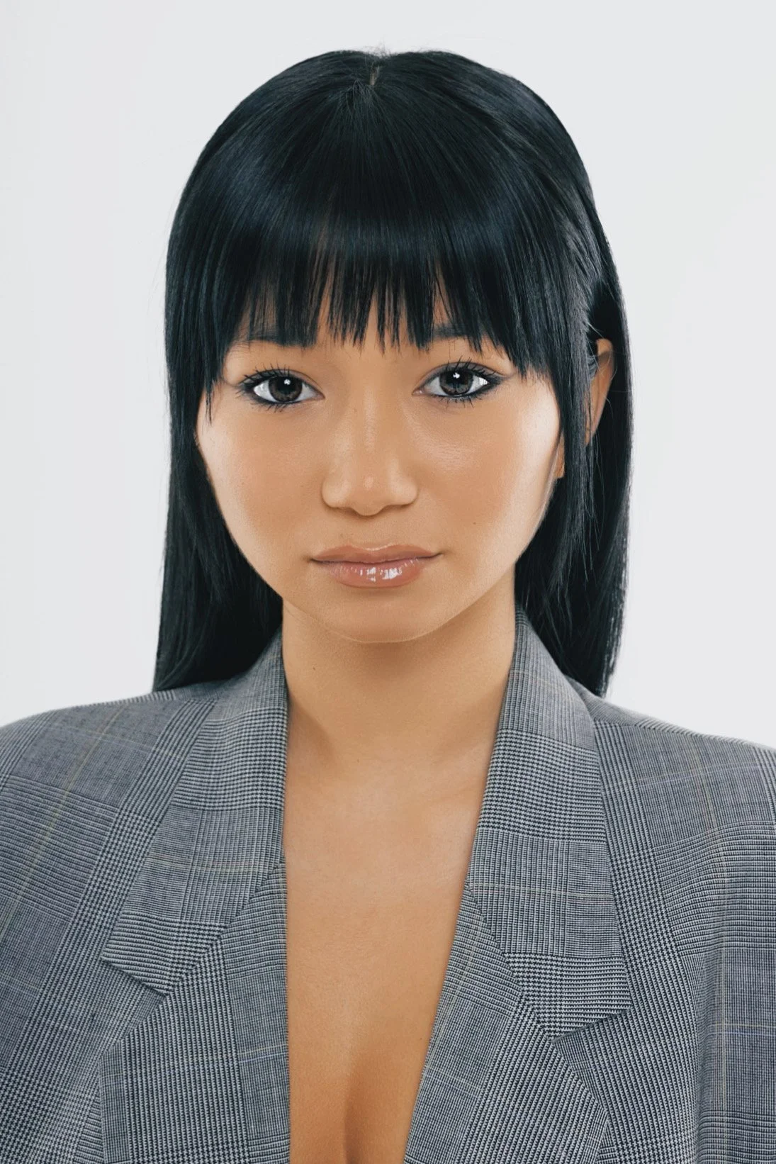 A woman with straight black hair, wearing a gray checkered blazer, facing the camera with a neutral expression.