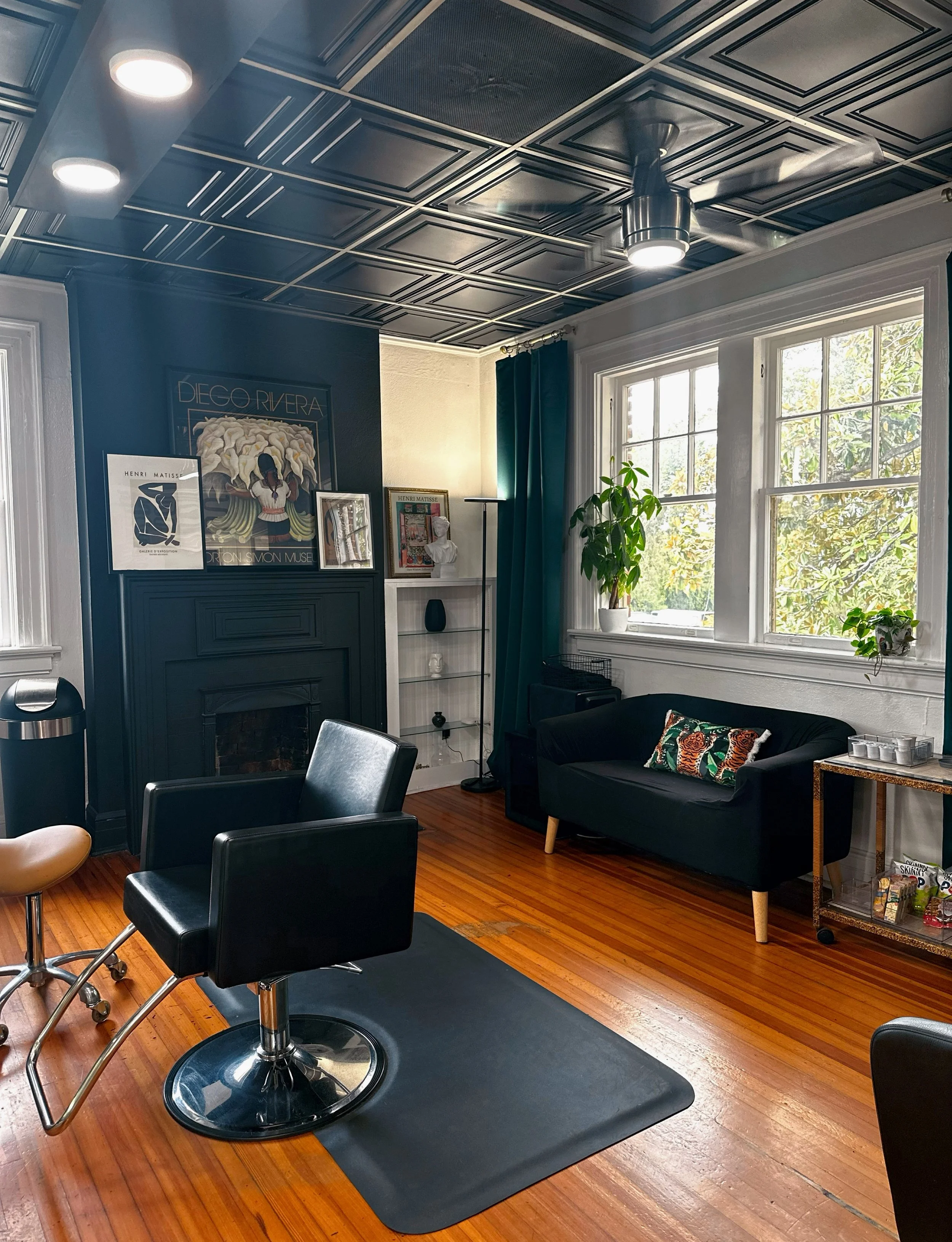 Interior of a room with a wooden floor, dark ceiling with a ceiling fan, a black fireplace with framed art on top, large window with green curtains, a black couch with colorful pillow, and a metal and glass side table with decorative items.