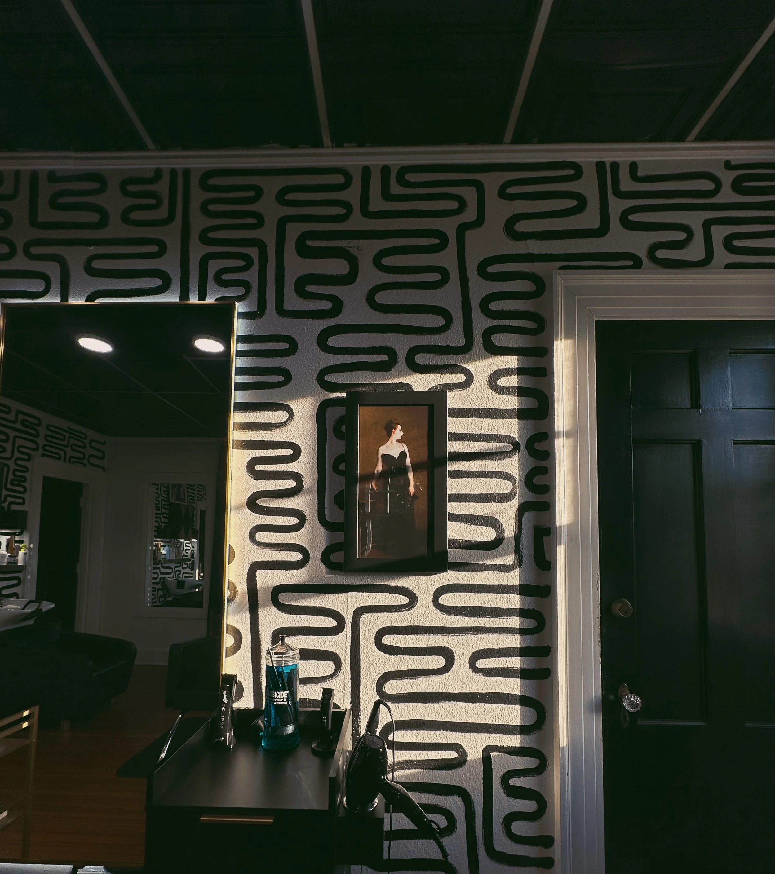 Interior of a salon with black and white patterned decor, a framed fashion photograph on the wall, a black counter with hair styling tools and a water bottle, and dark doors.