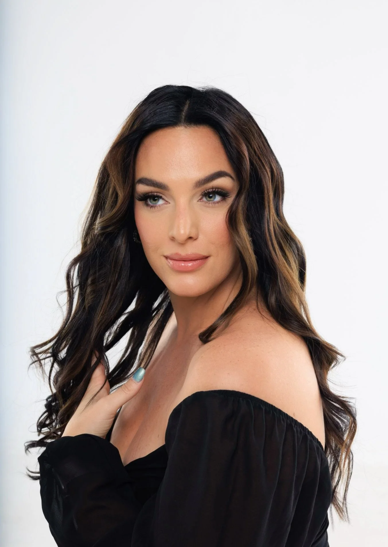 A woman with long wavy brown hair with blonde highlights, wearing a black off-the-shoulder top, touching her shoulder with her left hand, and looking over her shoulder against a plain white background.