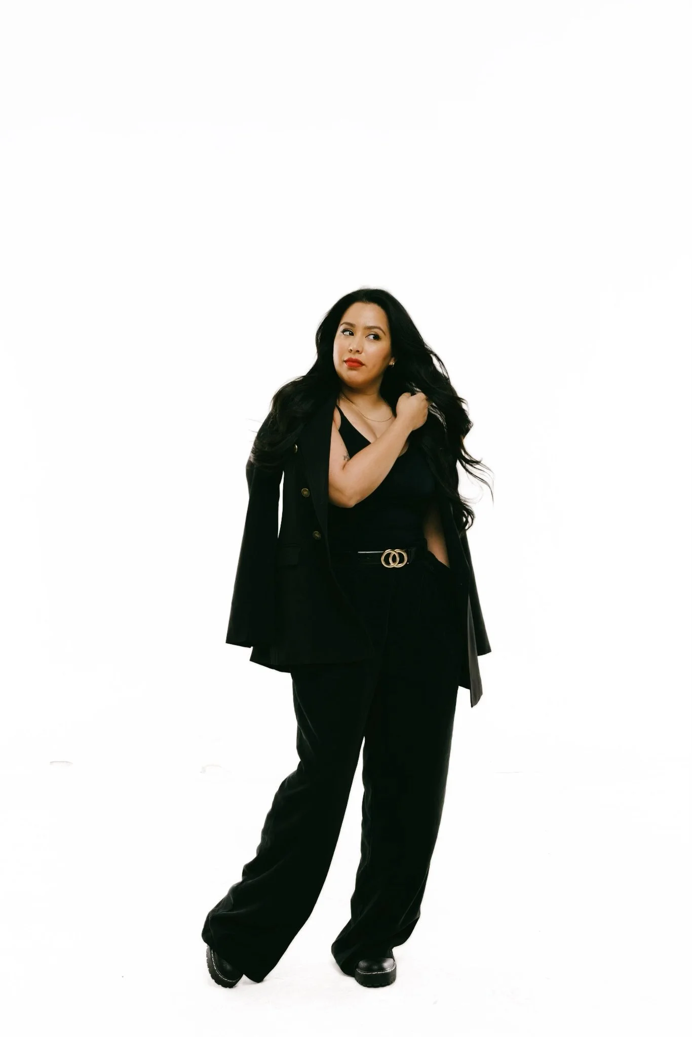 A woman with long black hair wearing a black outfit with a black blazer, high-waisted black pants, and black boots, standing against a plain white background, looking to her right with a confident expression.