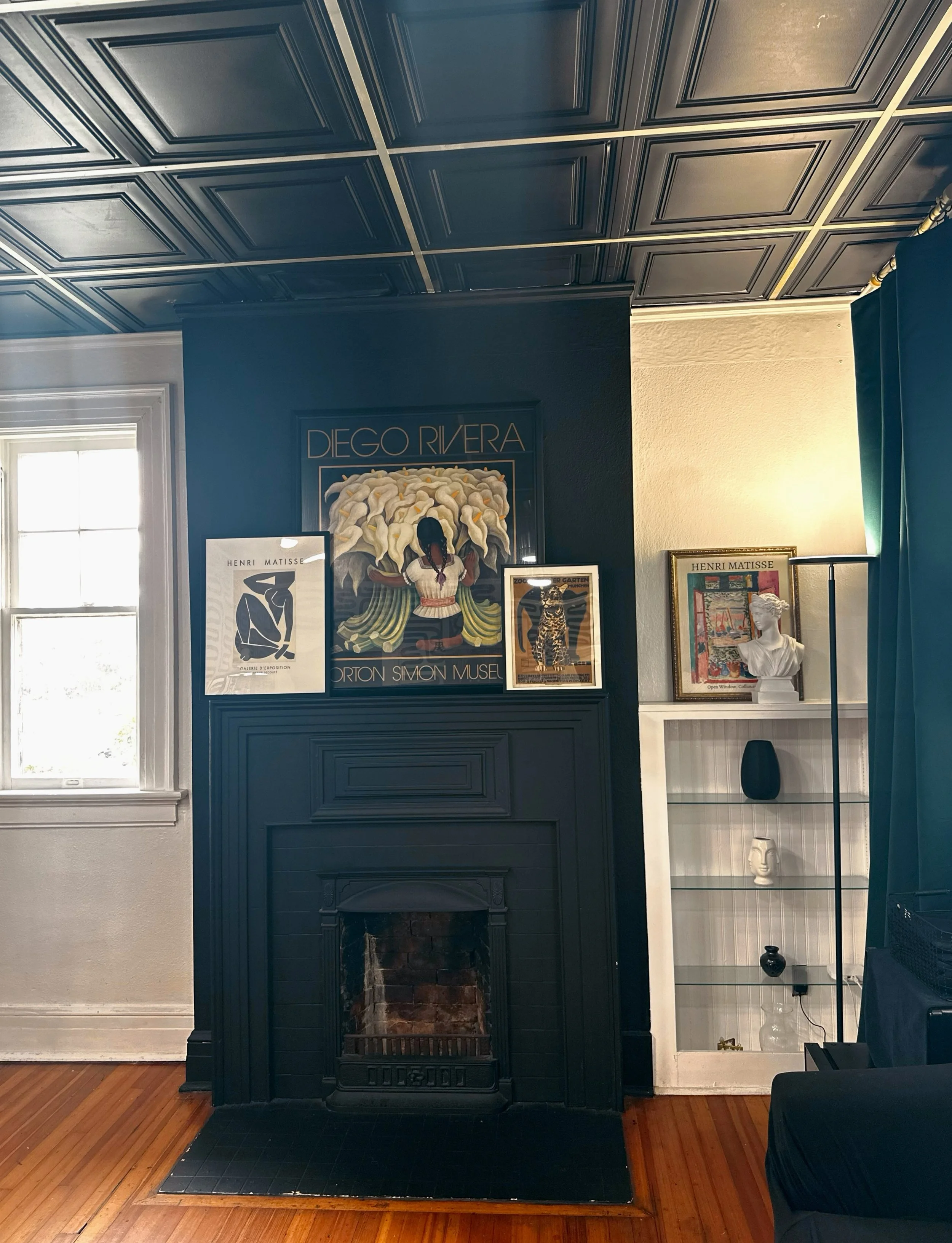 A room with a dark blue painted wall and a black fireplace with a mantel. Above the fireplace, there's a framed artwork titled 'DIEGO RIVERA' featuring a painting of a woman with white flowers around her. To the right of the fireplace, a white cabine