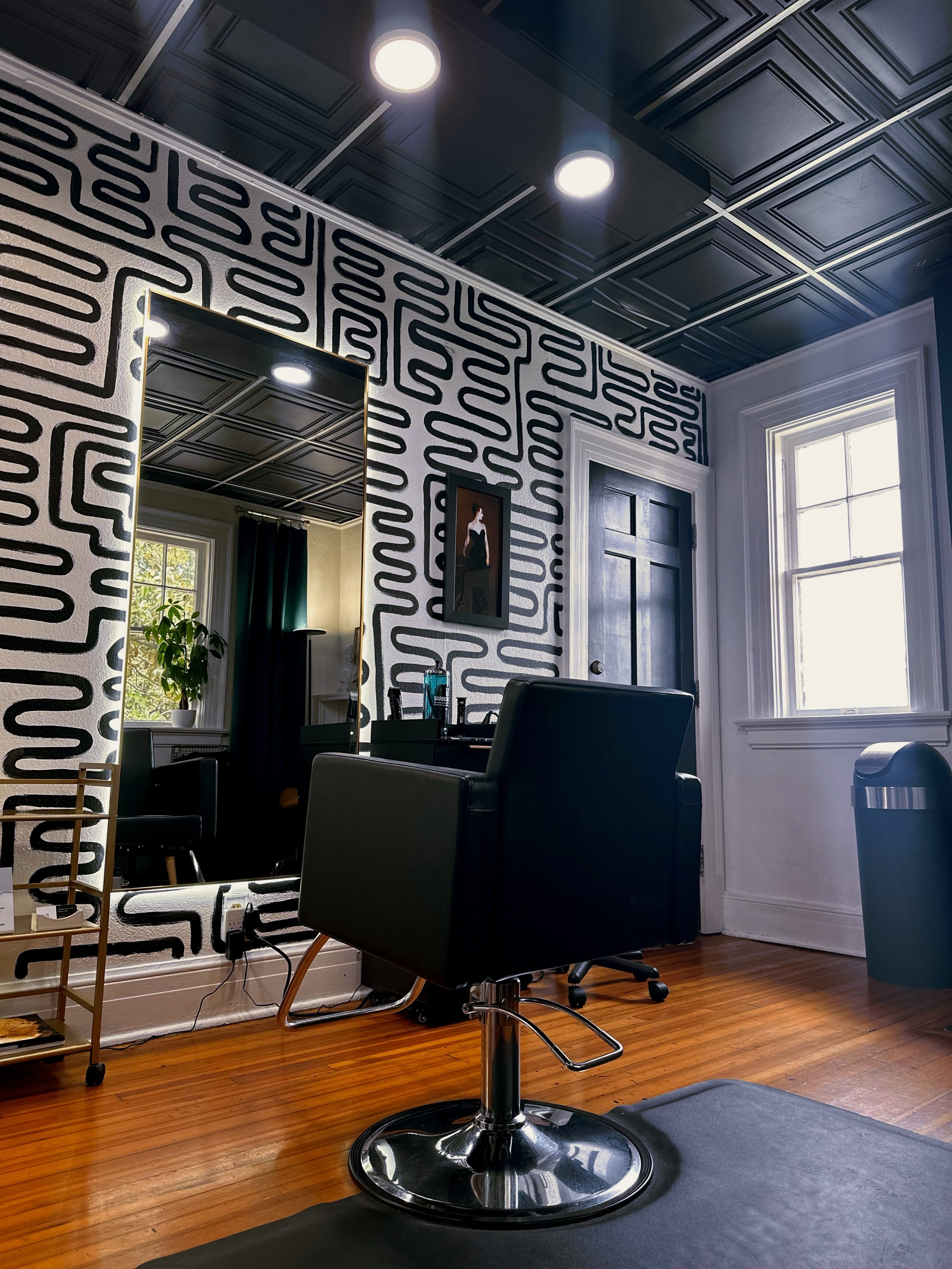 Interior of a room with dark ceiling tiles and a wall with black and white maze-patterned wallpaper, a window with white trim, black furniture including a chair, a mirror, and a small table. There is a water bottle on the table and a trash bin near t
