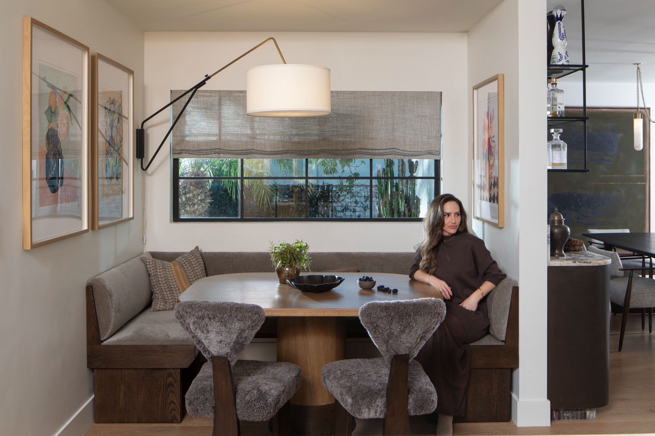 Woman sitting in a dining nook with upholstered bench and furry chairs, decorative artwork and hanging lamp, next to a kitchen with open shelving and a black table.