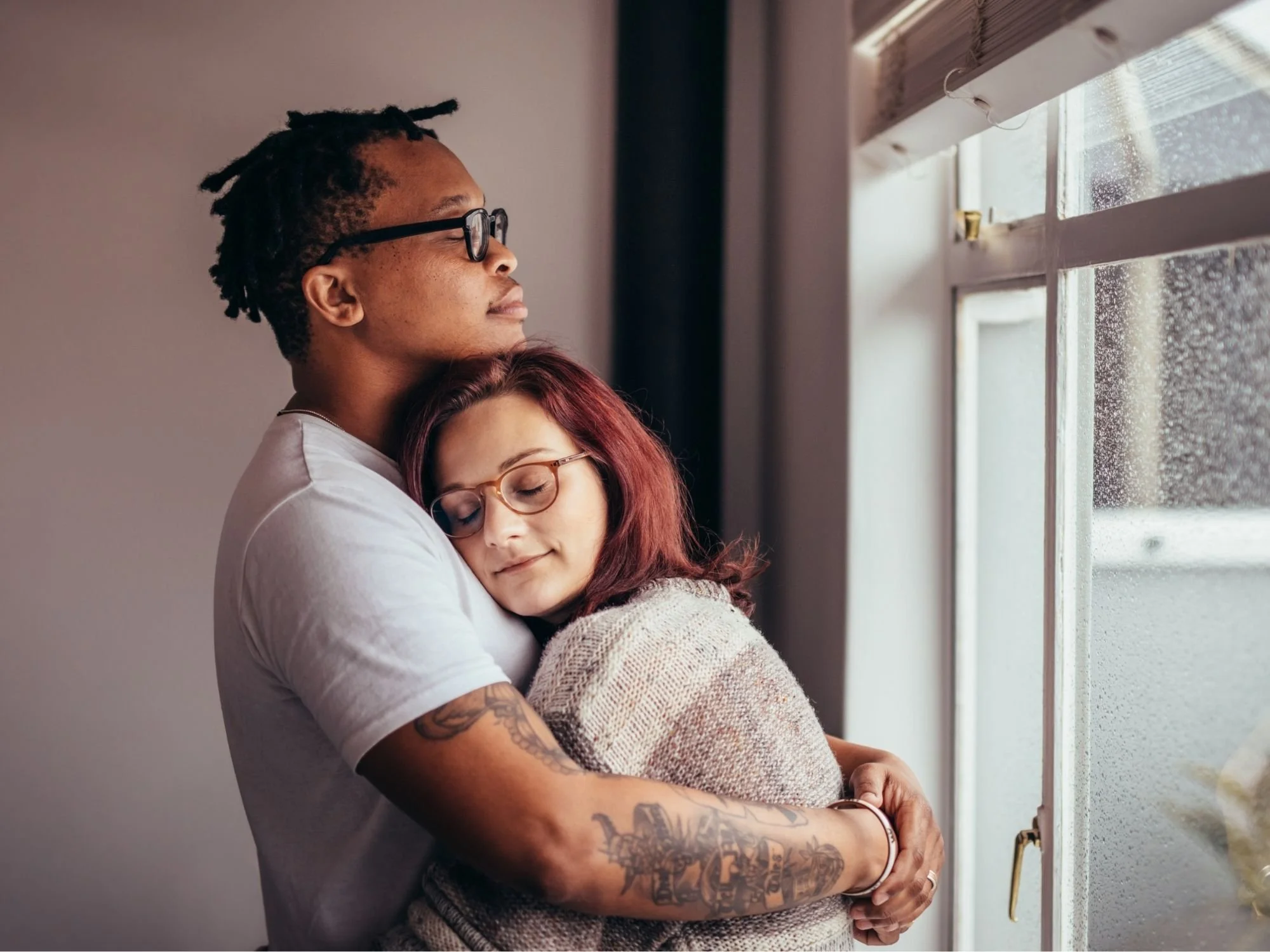 A man and a woman are hugging each other while looking out a window on a rainy day.