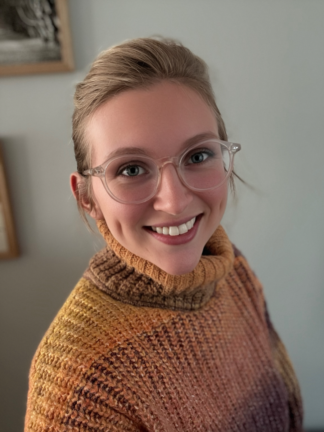 A woman with short blonde hair, wearing clear glasses and a multicolored knit sweater, smiling at the camera.