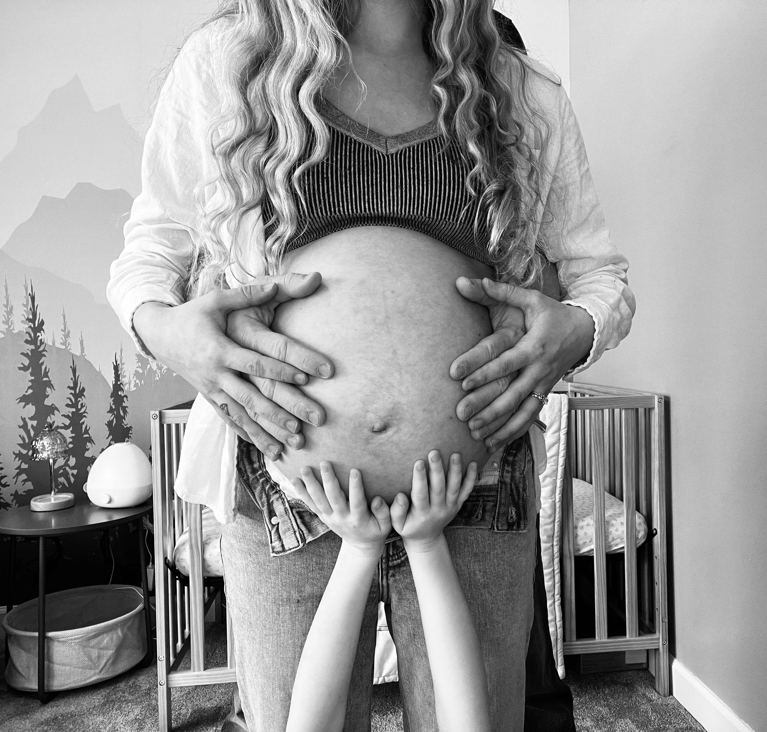A woman with long wavy hair is holding her pregnant belly with her hands. She is wearing a striped top and a jacket. The background includes a decorated wall with a mountain and trees painted on it, a small table with a lamp, and a wooden bed with a pillow.