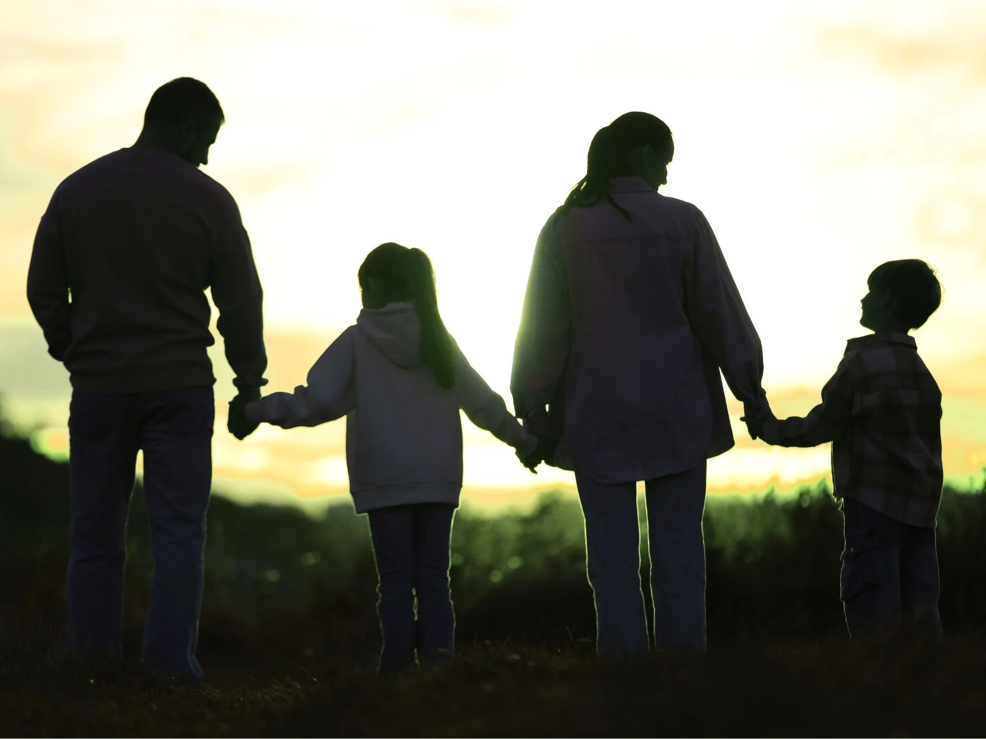 Silhouette of a family holding hands outdoors during sunset or sunrise with a bright sky in the background.
