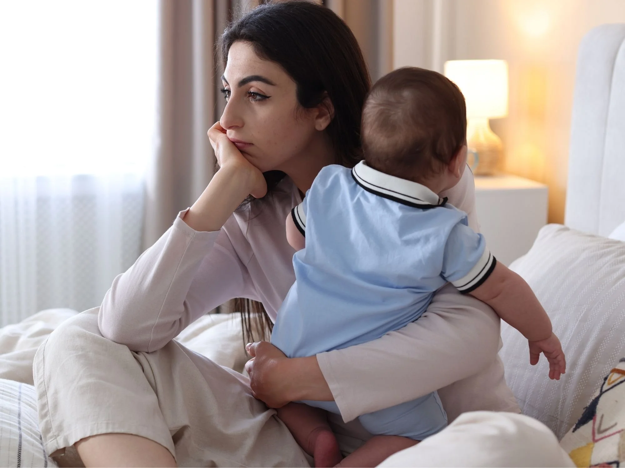 A woman with dark hair, wearing white pajamas, sitting on a bed with a baby in her lap. The woman rests her chin on her hand and looks away thoughtfully, while the baby, dressed in a light blue outfit with black trim, faces away from the camera. The room has warm lighting, a bedside lamp, and curtains in the background.